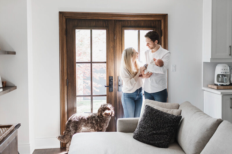 Mom and dad holding newborn son in their Toronto living room. They are standing by the patio doors and are smiling lovingly at each other.