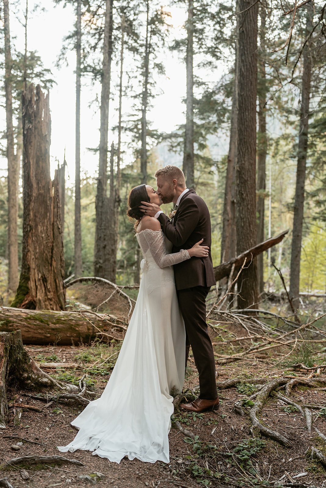 A bride and groom share a kiss surrounded by tall pine trees and golden light filtering through the forest in Glacier National Park, captured by Sydney Breann Photography.