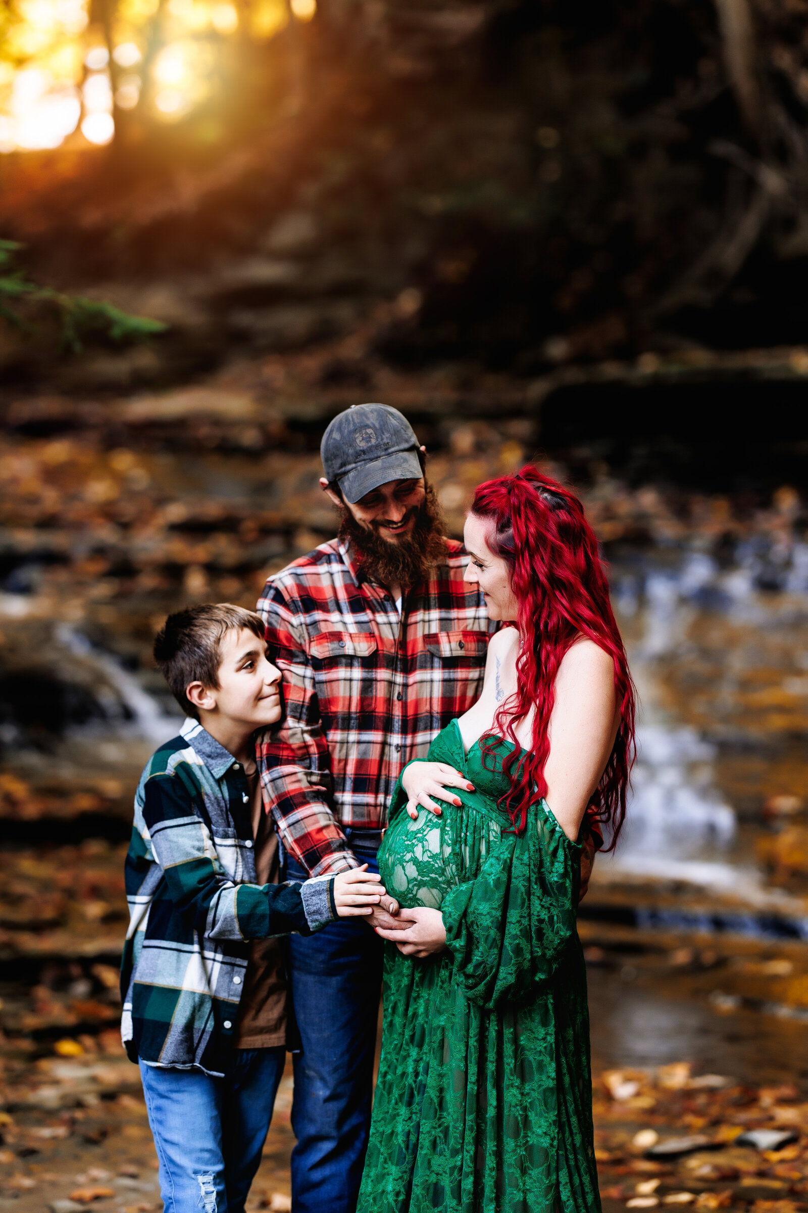 Family of three snuggles up together holding mothers pregnant belly in front a beautiful waterfall with fall foliage