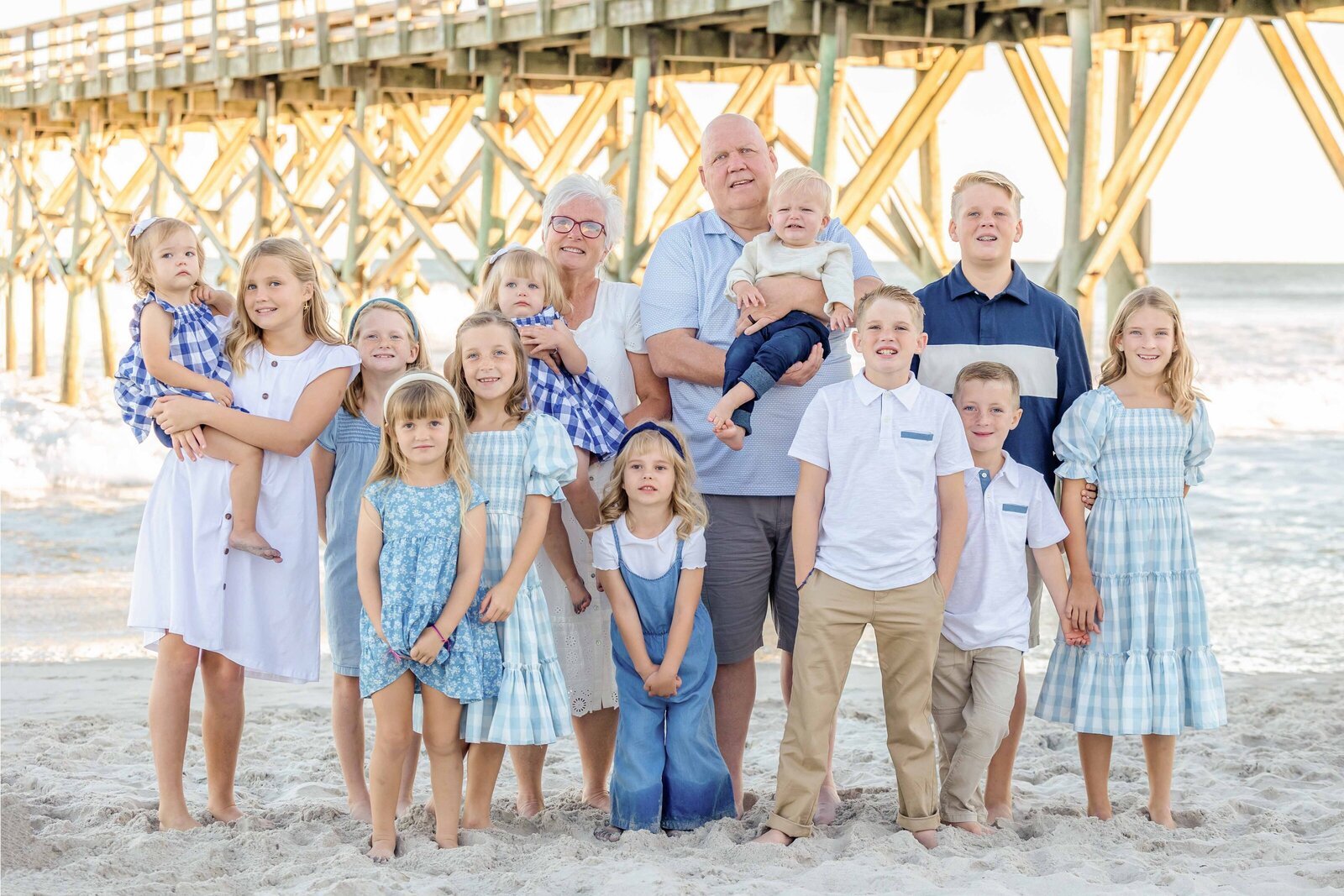 A multi-generational family stands under a pier, dressed in coordinated blue outfits. The ocean waves and wooden beams create a dynamic setting, ideal for destination family photographer NC, highlighting joyful family connections.