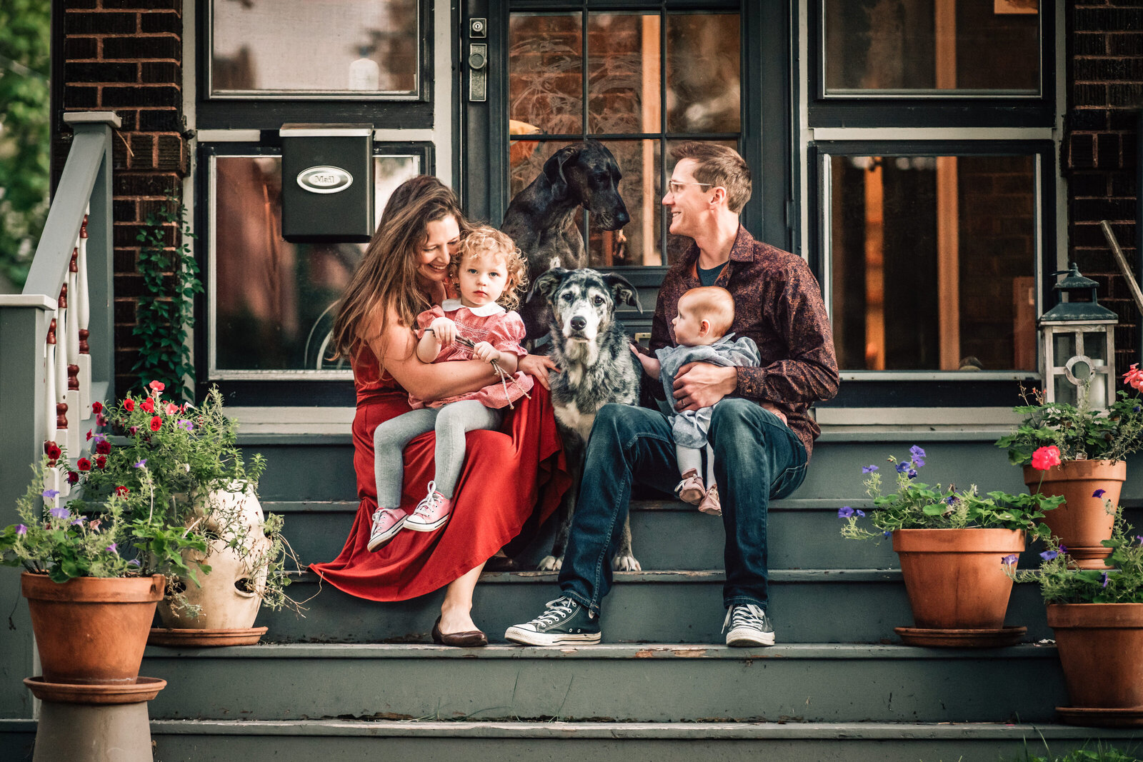 A cozy family scene on the front steps of a home, with the parents seated and holding their two young children, surrounded by flower pots and their two dogs.
