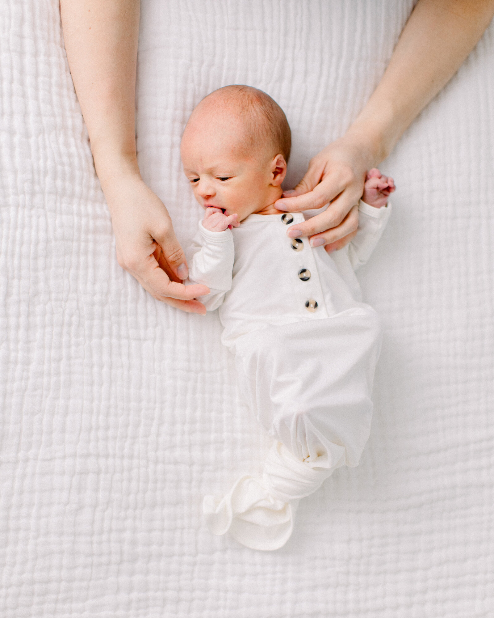 Baby in white Lou Lou & Co knotted onesie with is mother's hands soothing him on a white muslin blanket by NH newborn photographer Fieldstone Studio.