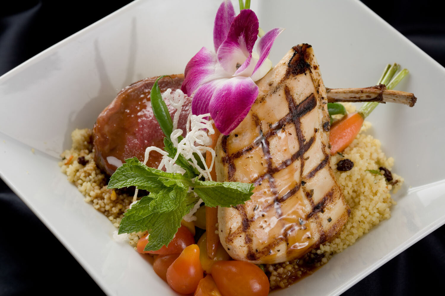 Grilled pork chop dish garnished with couscous, vegetables, and a purple orchid