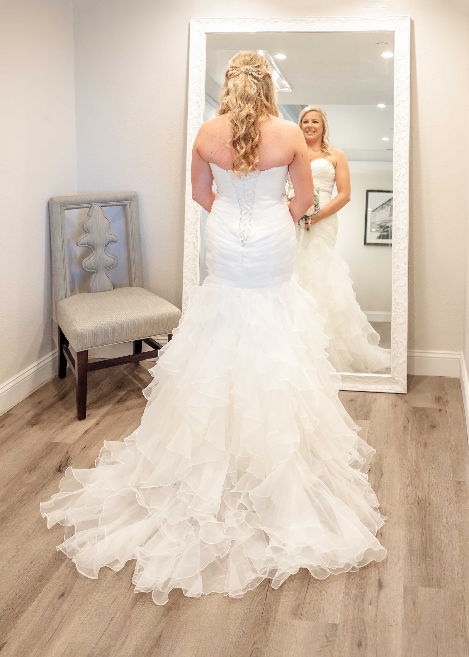 A bride in a layered, ruffled wedding gown admires her reflection in a full-length mirror in an elegant bridal suite, captured during a Wilmington, NC wedding, highlighting authentic moments and bridal preparation.