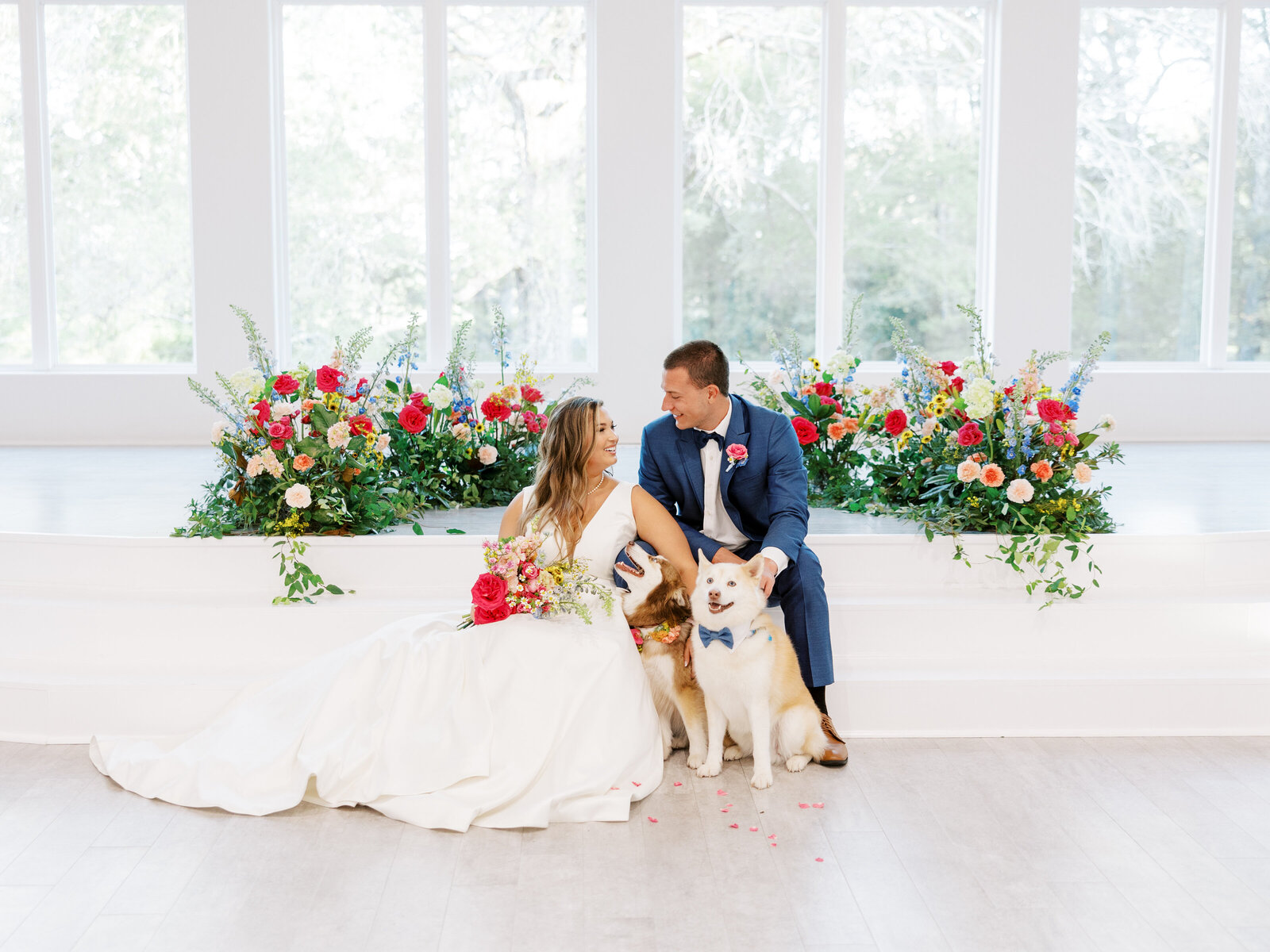 bride and groom sitting together after ceremony with their two dogs surrounded by lush pink, blue, yellow, and white florals