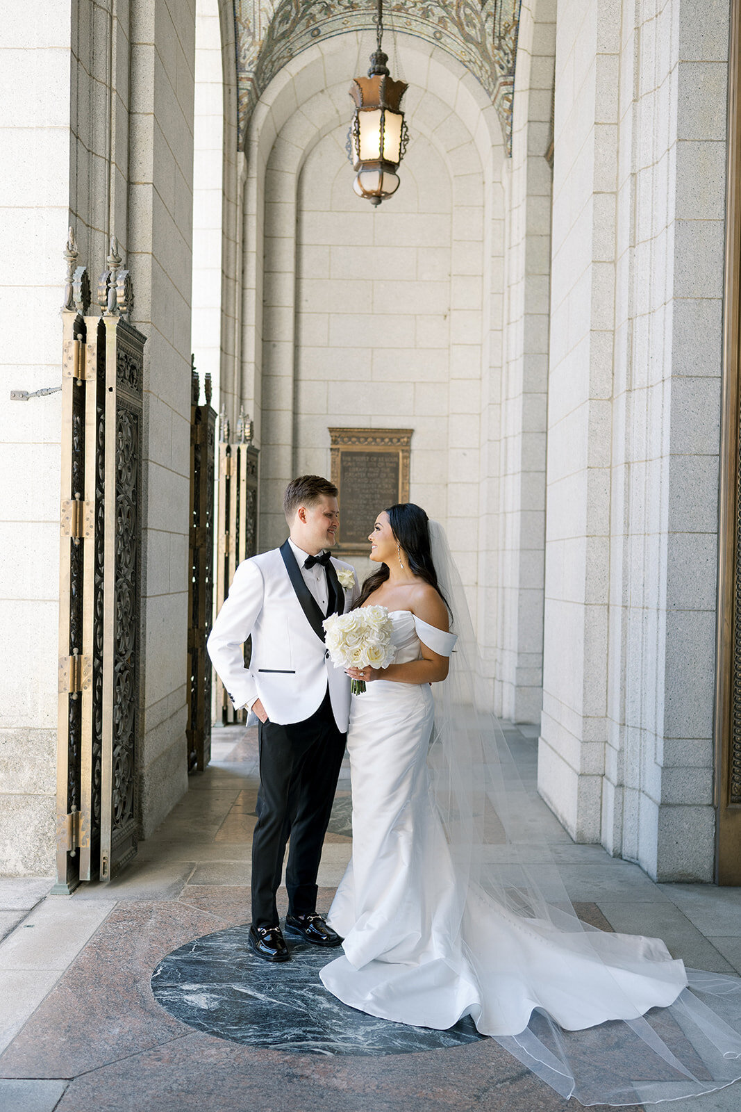 Elegant bride and groom share a romantic moment beneath ornate stone archways at a timeless historic St. Louis library — bride in classic off-the-shoulder gown holding white roses, groom in white tuxedo jacket. Captured by Jordan Grover Photo, a Missouri-based traveling wedding photographer known for refined, documentary-style wedding photography.