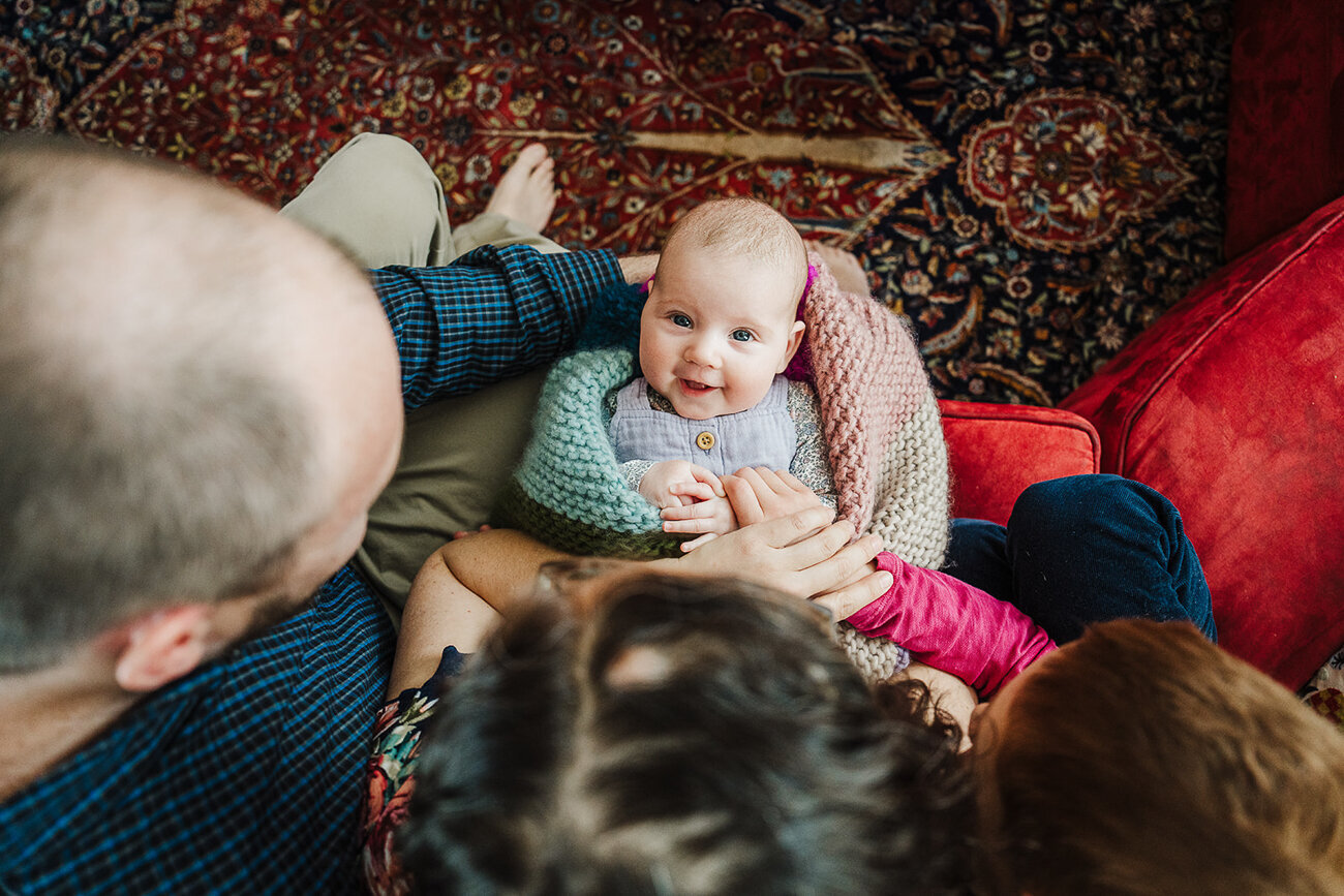older baby looks straight at camera while wrapped in rainbow blanket in parents laps