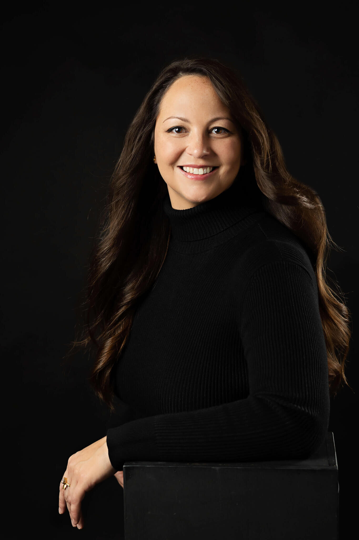 young professional posing  for her professional headshots, she is wearing a black turtleneck on a black backdrop with a big smile