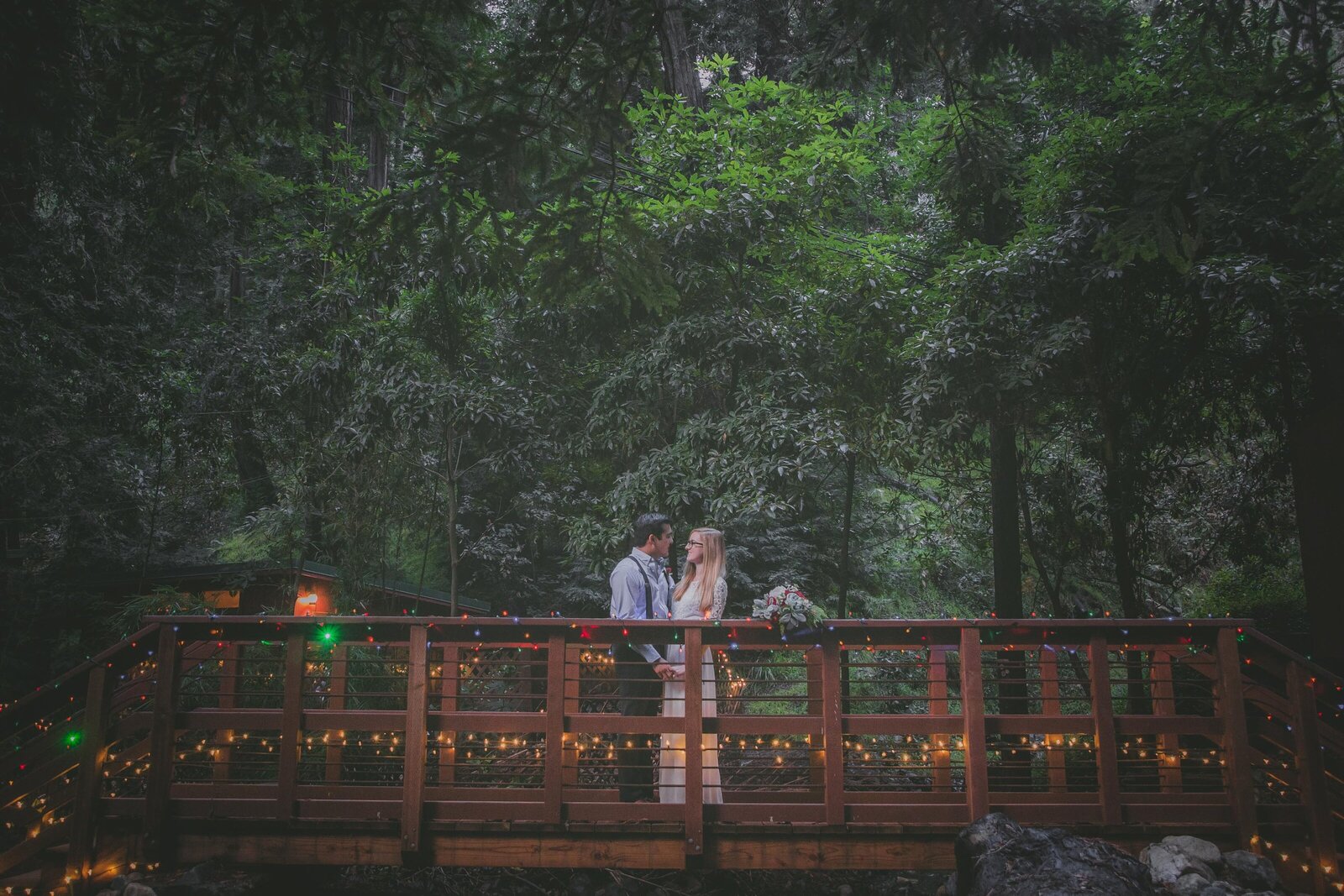 Couple looks at each other on bridge surrounded by redwood trees.
