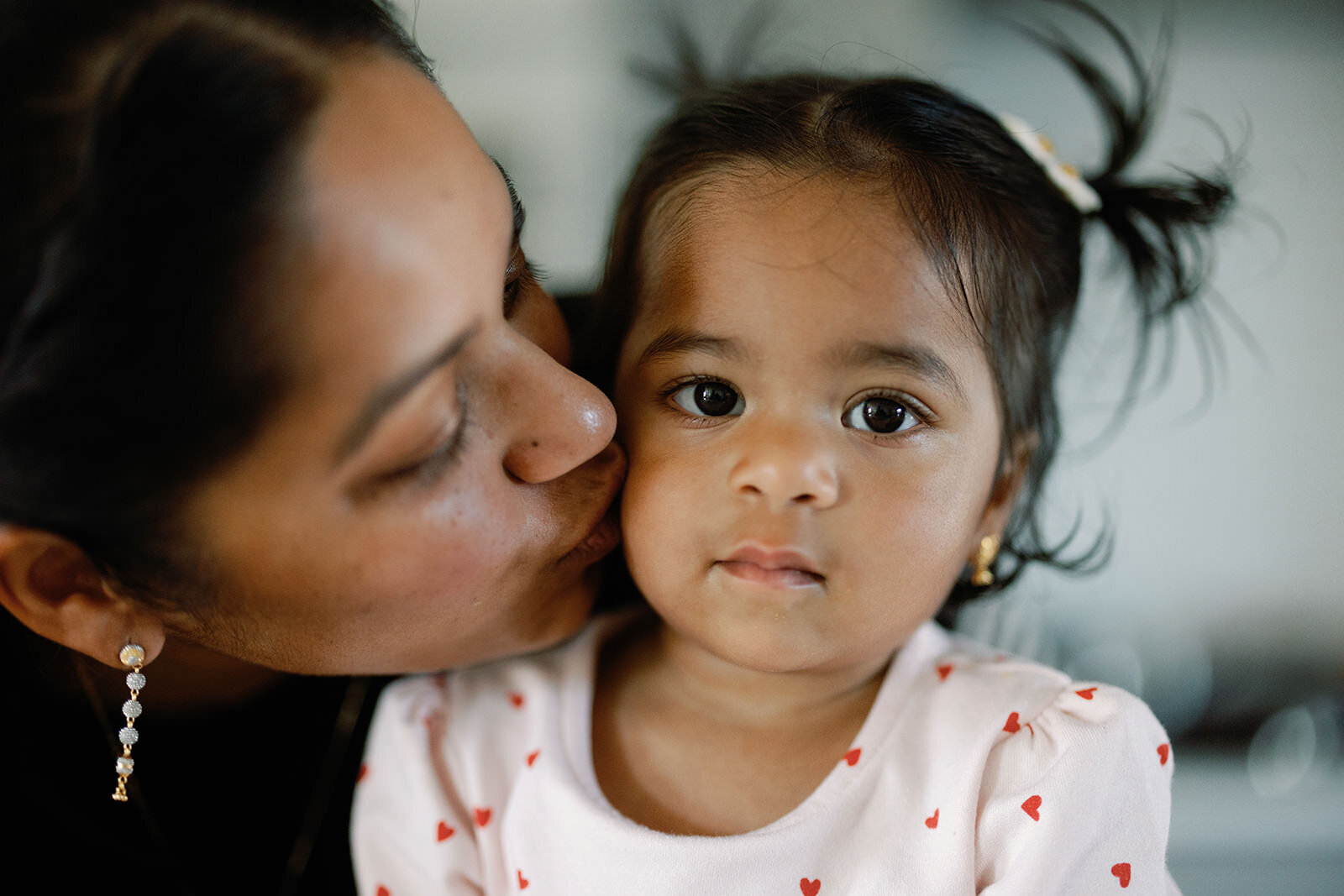 A touching moment of a mother gently kissing her young daughter on the cheek, with the child gazing softly at the camera.