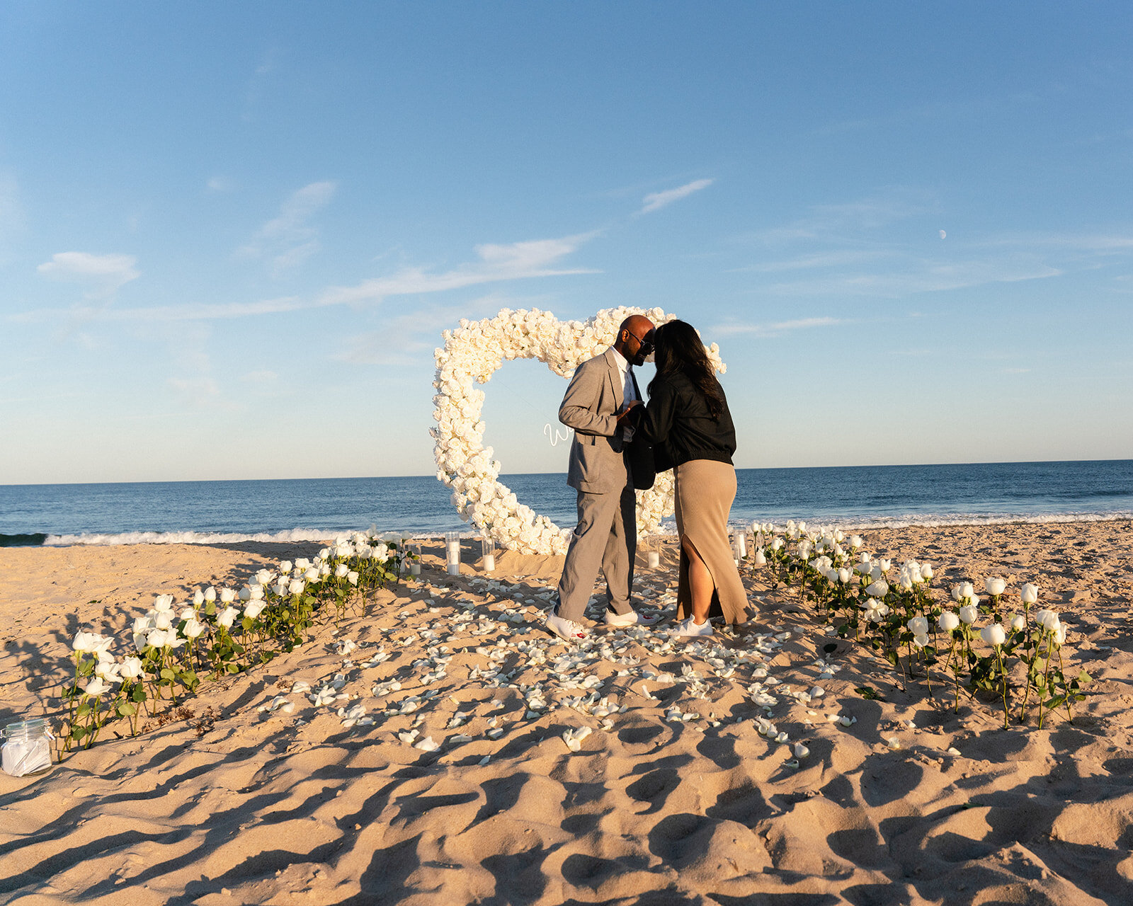 Romantic beach proposal with a white floral heart arch and petal-lined aisle, designed by Yours Truly, a wedding planner and designer in New Jersey.
