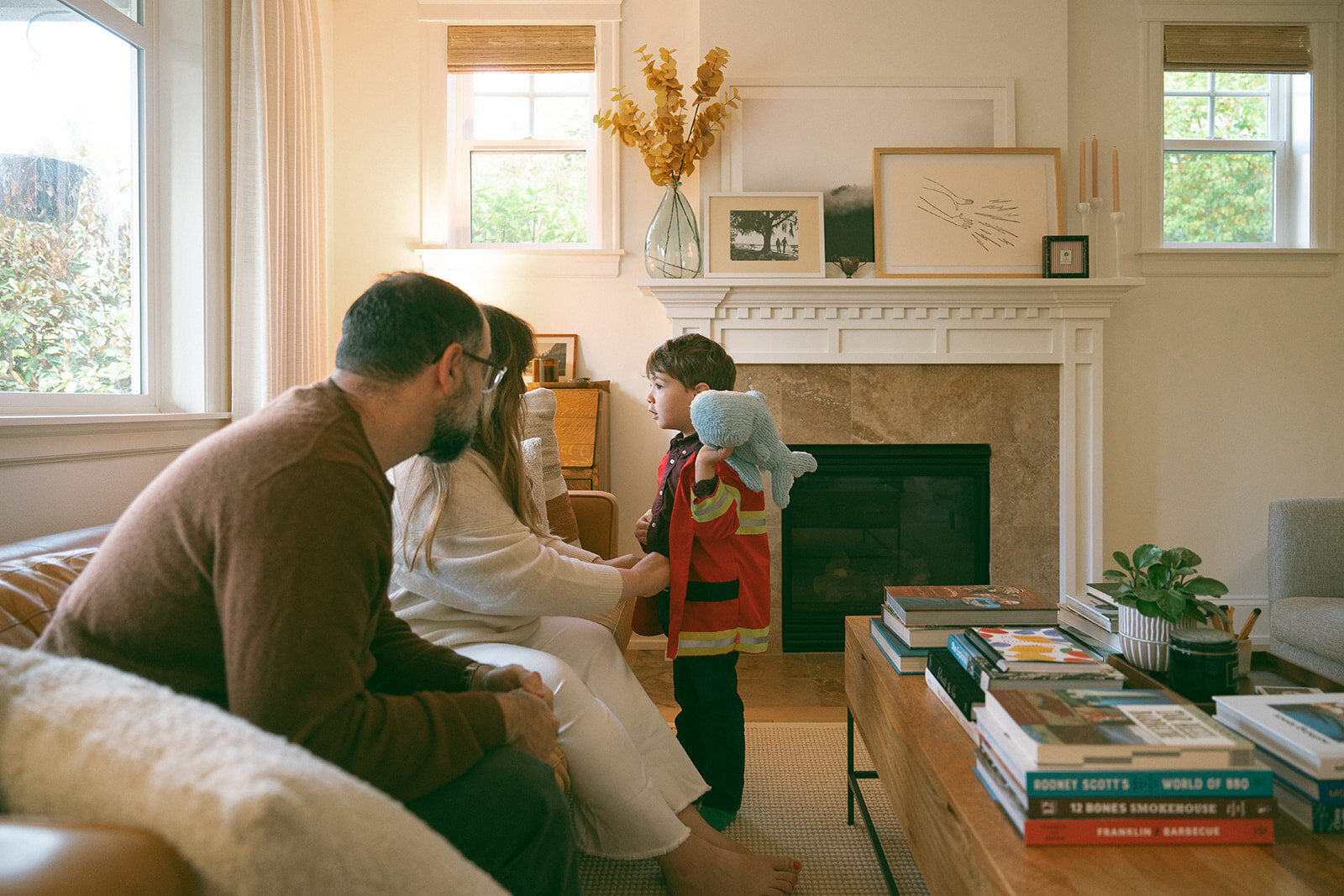 Parents with their young son in a firefighter jacket standing by the fireplace during candid at-home family photography in Orange County, California.