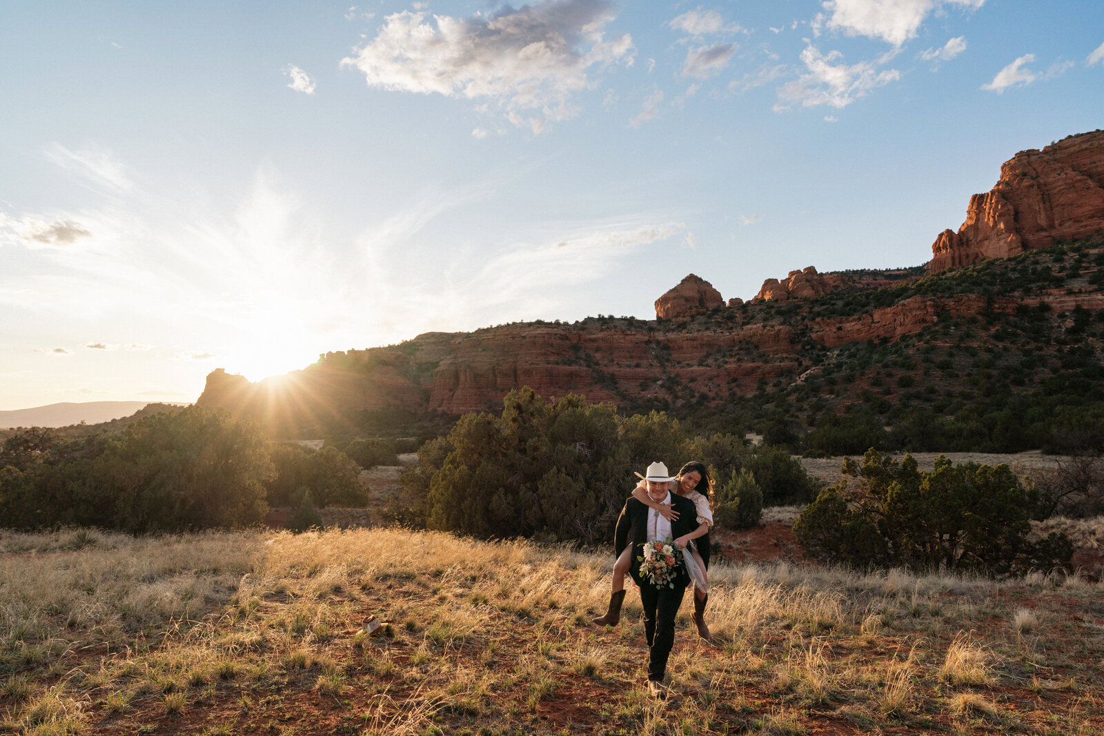 Bride in cowboy boots riding piggyback on groom wearing a cowboy hat during their western-inspired elopement at Bear Mountain in Sedona, Arizona, with golden hour sunlight and red rock cliffs in the background.