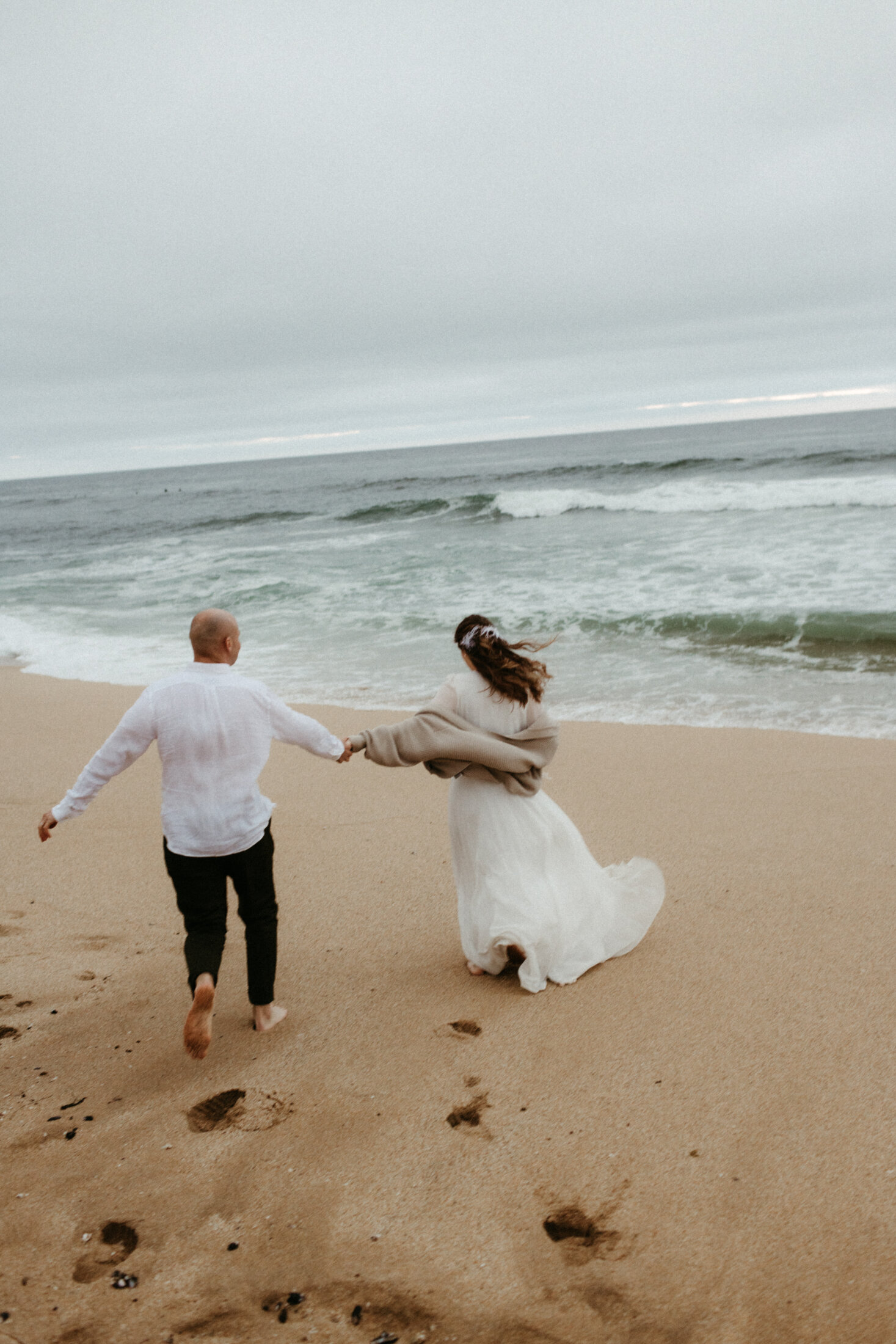 A joyful couple holds hands while running along a sandy beach towards the ocean in San Francisco, the woman's white dress billowing behind her on a cloudy day.