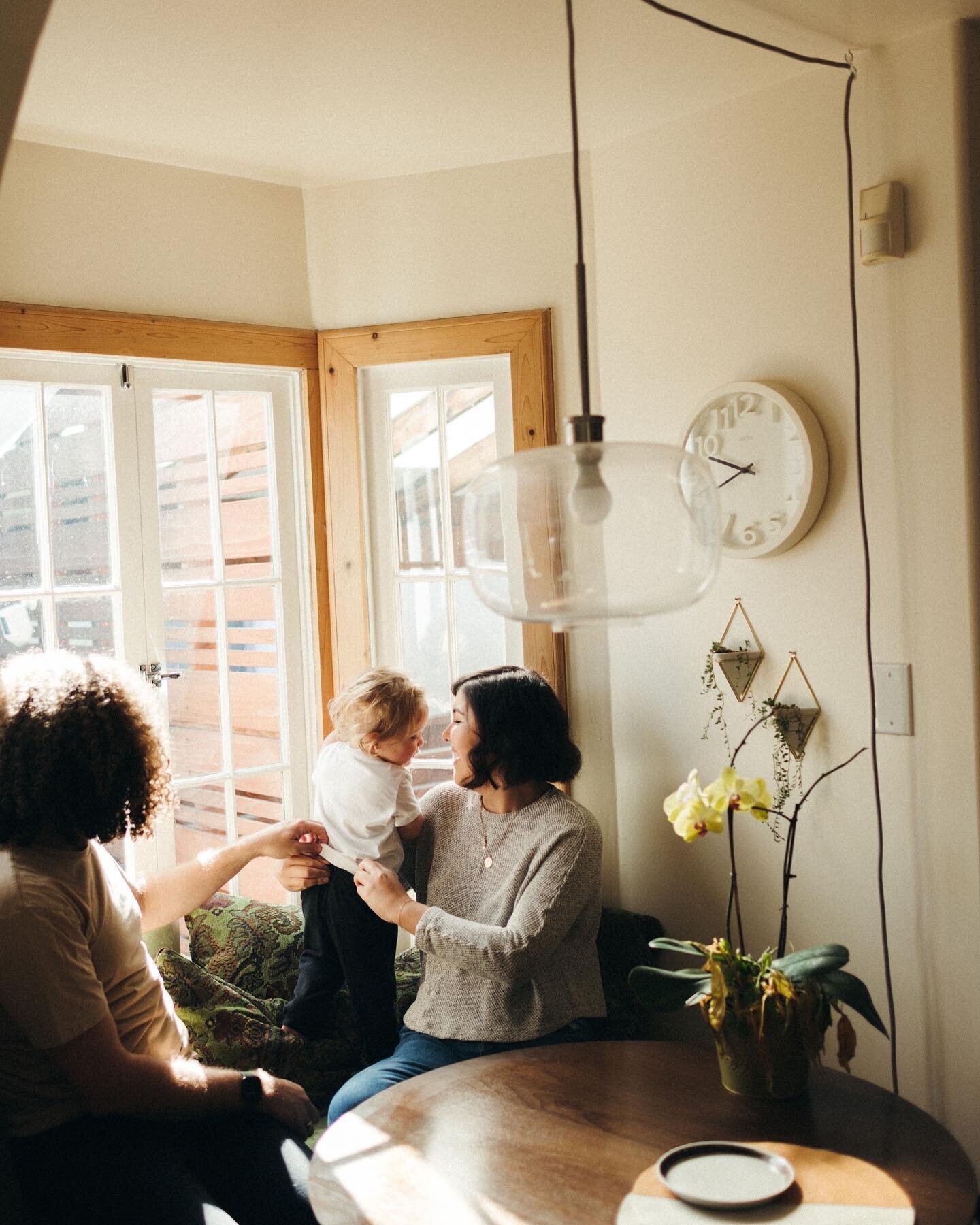 A cozy family moment in a sunlit room, featuring two adults and a child near a wooden table, with touches of greenery and a modern lamp hanging above.