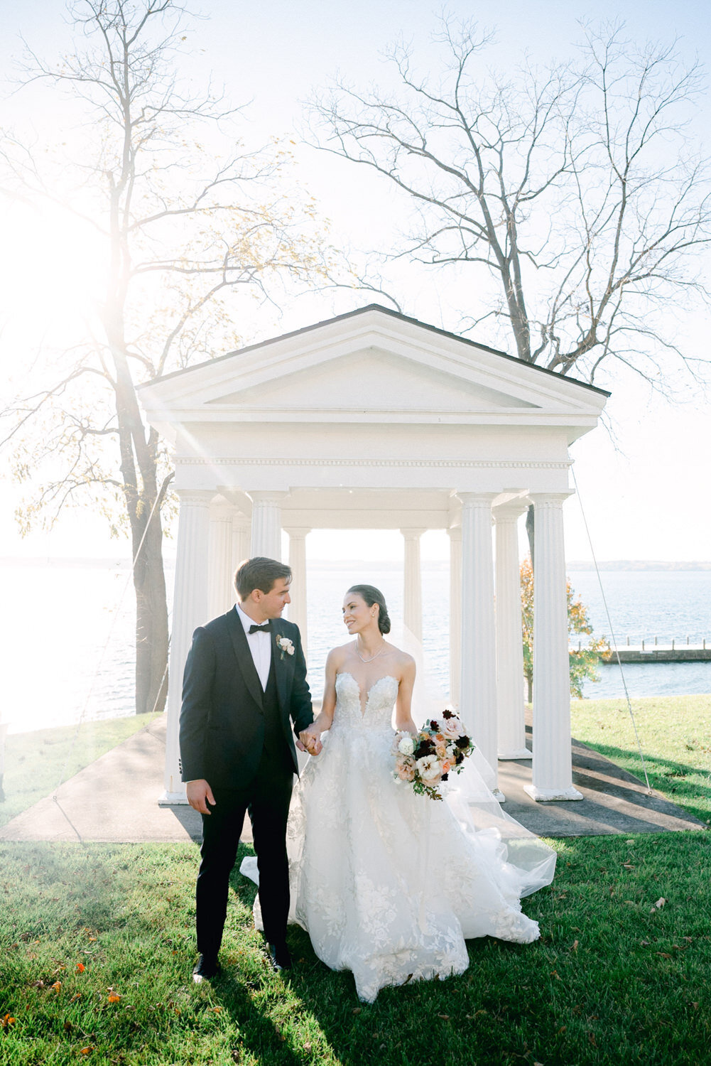 Bride and groom walk in front of white columned pergola.