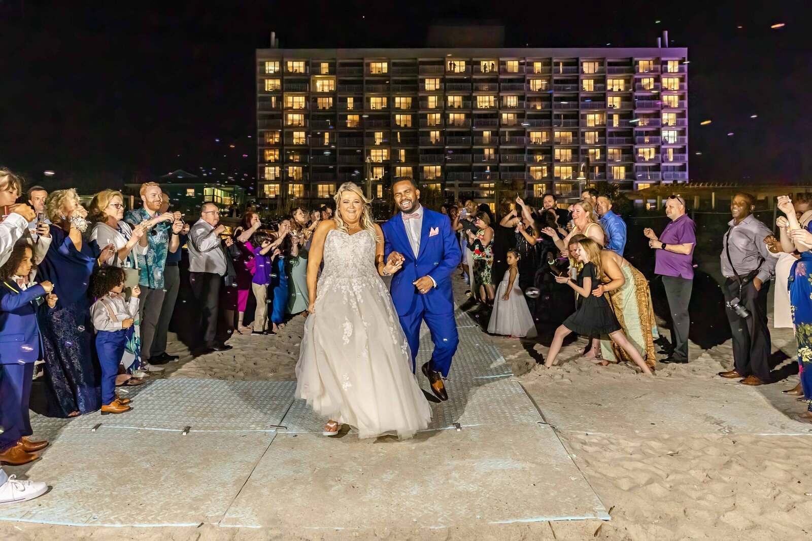 The bride and groom walk hand-in-hand during a sparkler exit on the beach at a Carolina Beach, NC wedding, surrounded by cheering guests and glowing coastal lights.