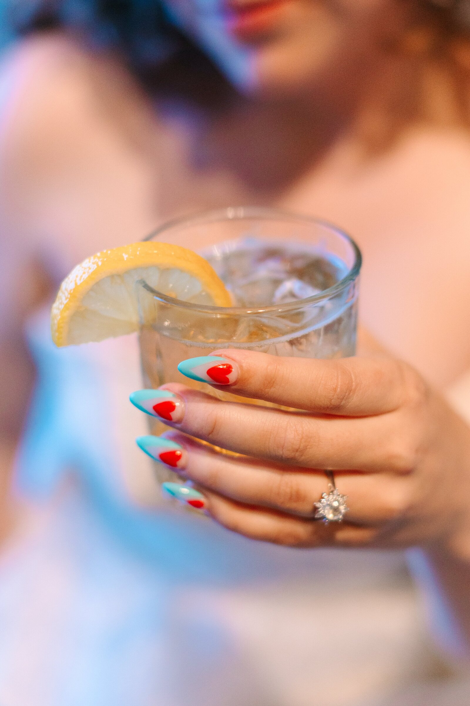 A closeup of a bride's hand holding a cocktail showcasing a starburst diamond ring and retro painted fingernails with turquoise tips and red hearts