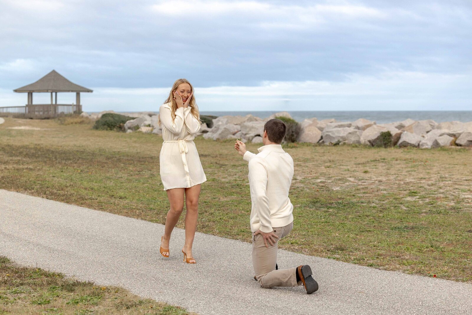 A romantic beachside proposal as a man kneels on one knee holding out a ring, while the woman, dressed in a white outfit, covers her face in joyous surprise. The ocean and a gazebo are visible in the background, creating a picturesque setting for North Carolina wedding photography.  