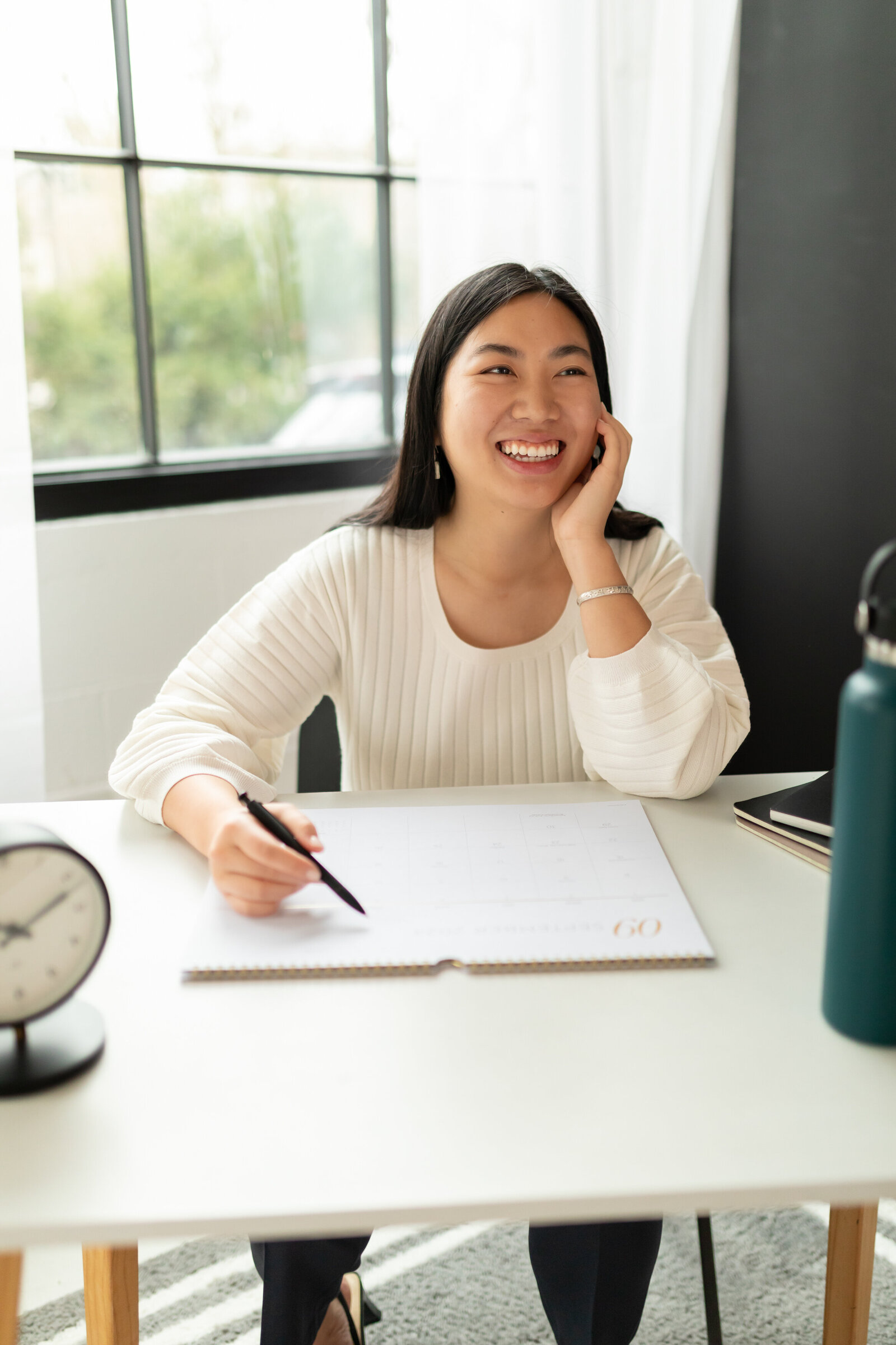 woman sits at desk in front of industrial windows while holding pen to paper desk calendar and smiles off camera with face resting in her hand