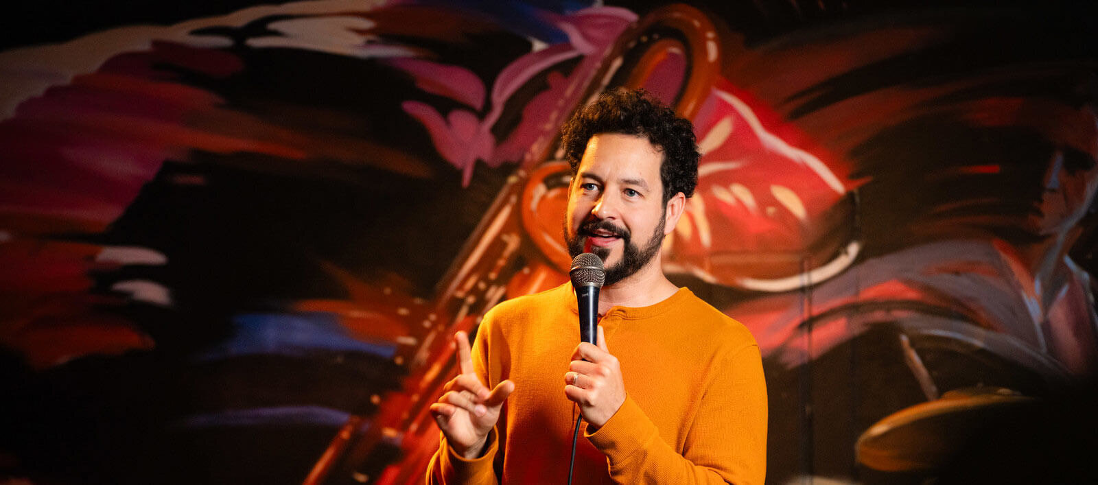 Comedian, Ahmed Bharoocha, holding a microphone and standing in front of a red background