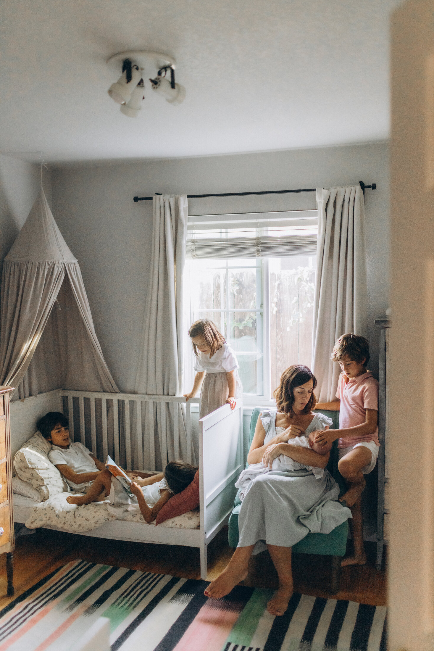 A cozy children's bedroom with one child in each of two bunk beds and a woman reading to two children sitting beside her. the room has curtains, a striped rug, and soft lighting.