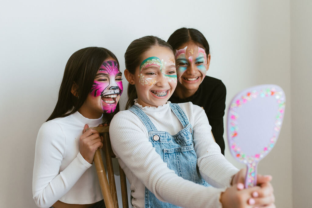 three girls with painted faces looking in to a mirror