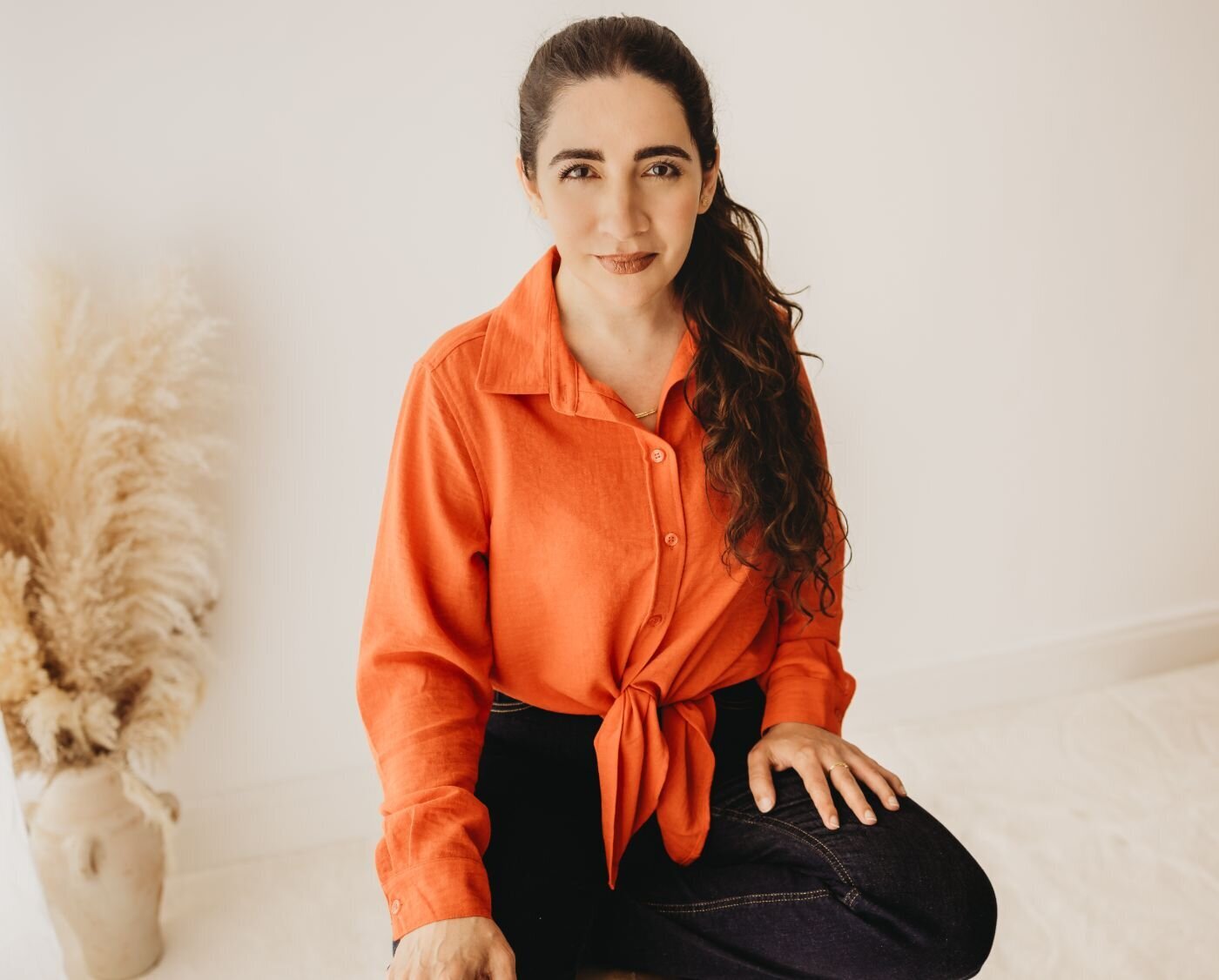 A woman with long, dark curly hair, wearing an orange shirt tied at the waist and dark jeans, smiles at the camera. Pampas grass in a vase is to her left