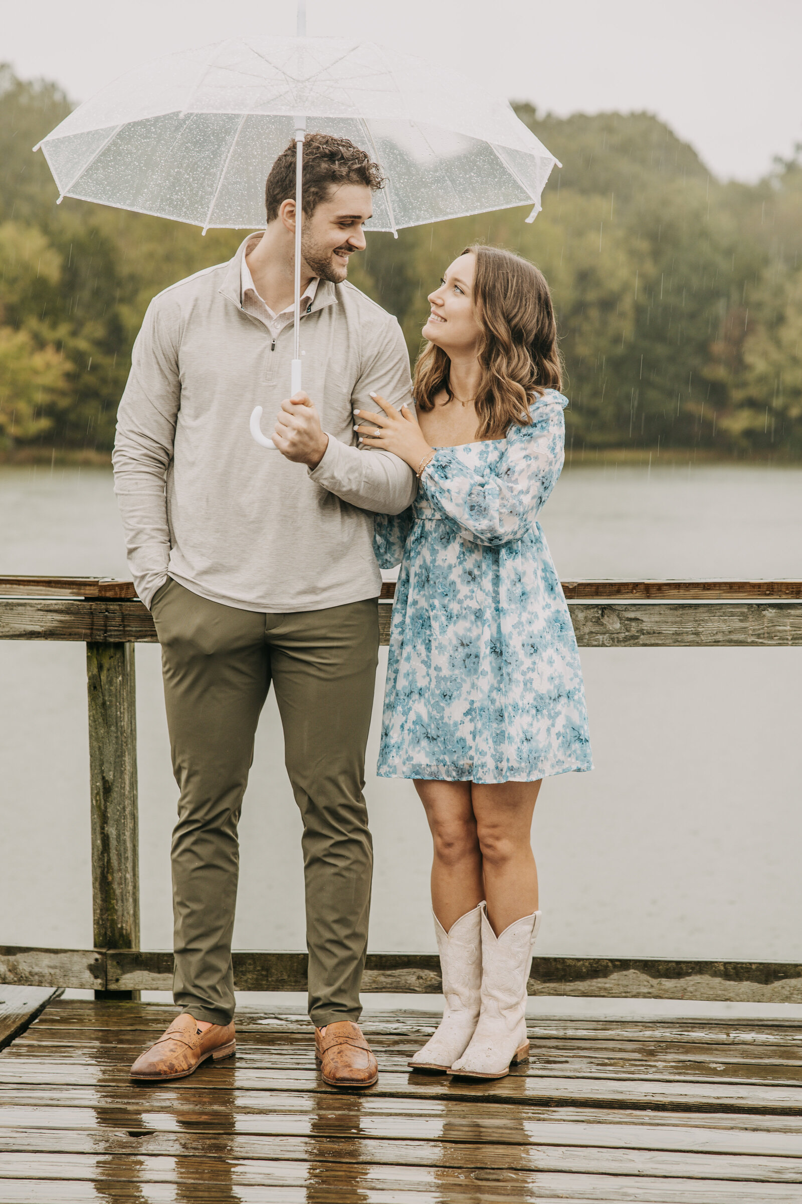 Drake and Lauren smiling at each other, standing under an umbrella on a rainy day during their engagement session at Shelby Farms Park.