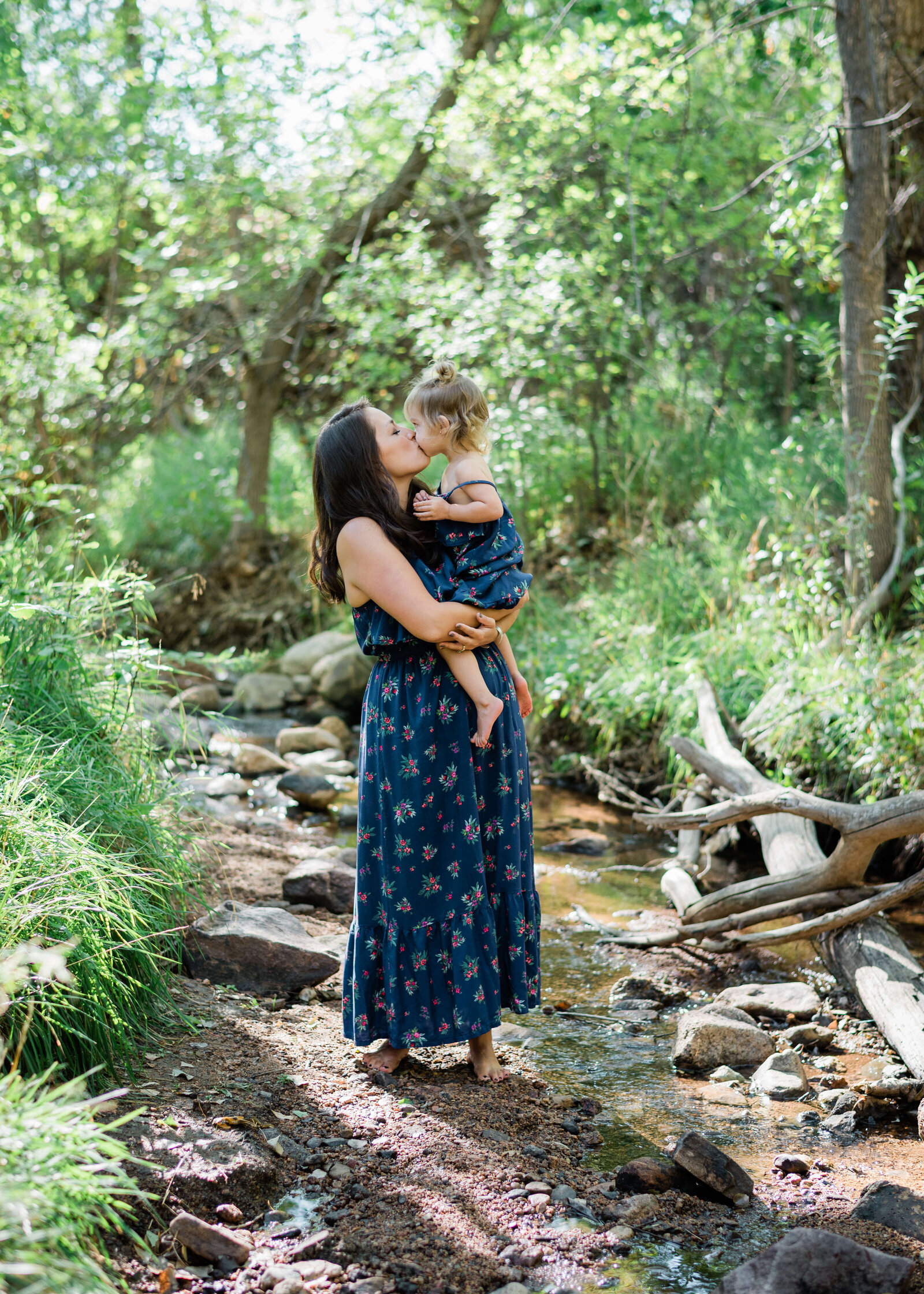 Barefoot and carefree, a mother holds her toddler and kisses her while standing in a stream