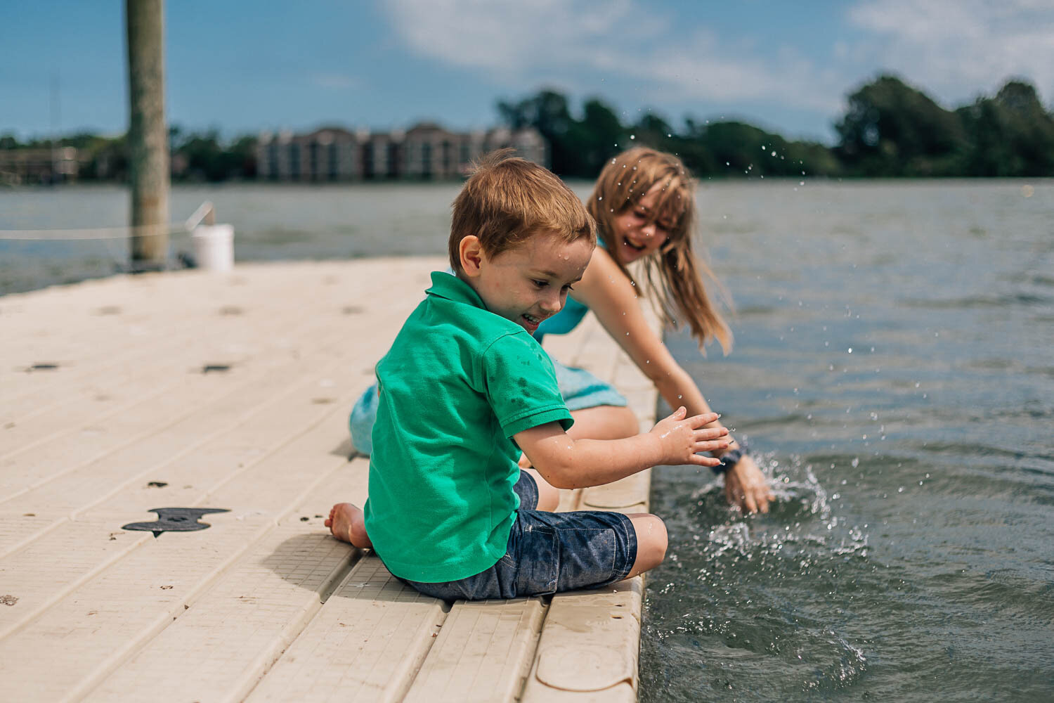 brother and sister sit on a dock together in Hampton Roads Virginia and splash water at one another and laugh, a candid moment captured by the best family photographers in Hampton Roads