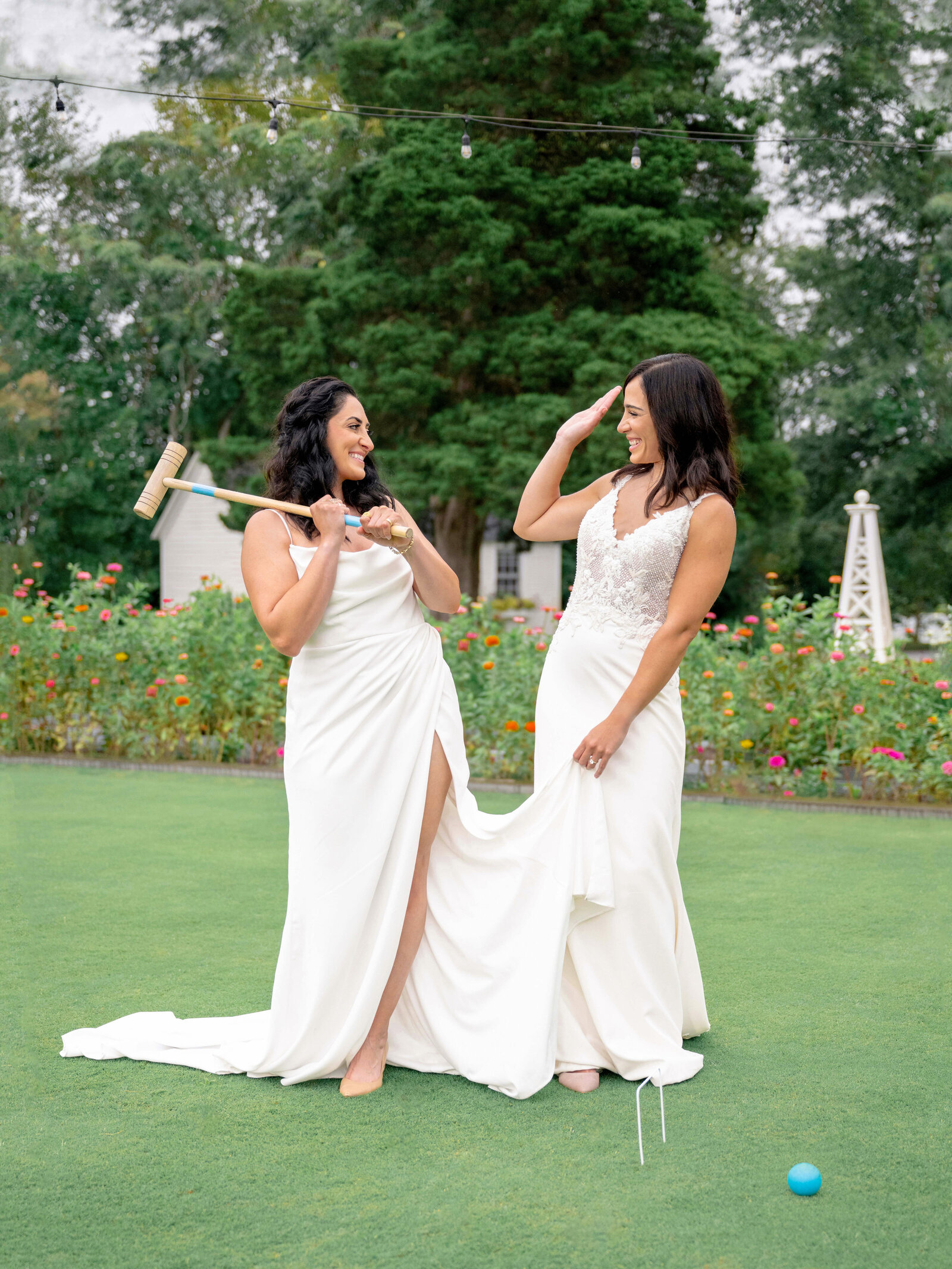 Two women in elegant white dresses stand on a grassy lawn. One holds a croquet mallet playfully, while the other lifts her dress slightly. Both are smiling and surrounded by a garden with blooming flowers.