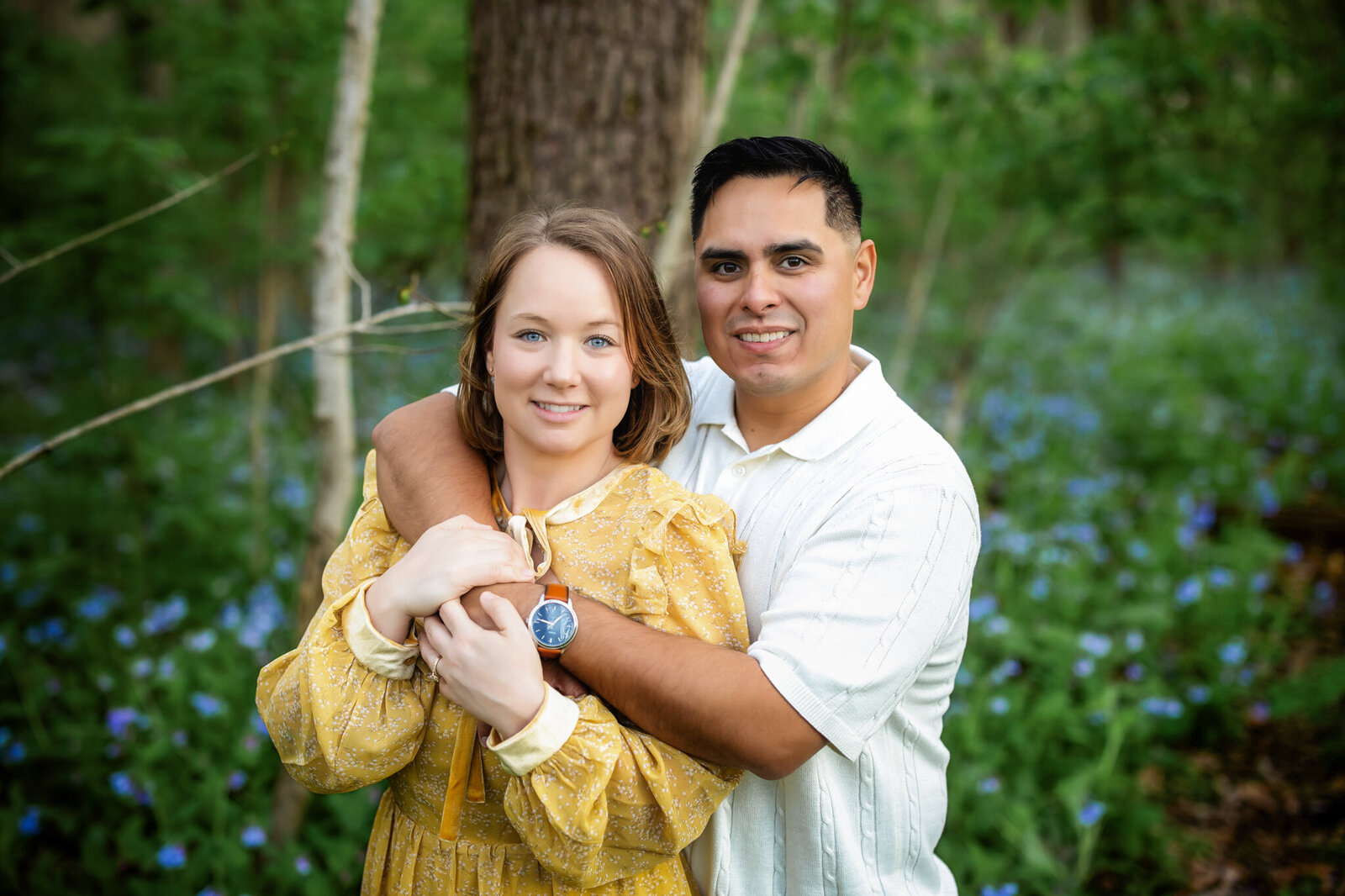 A man and woman stand close together in a forest, smiling at the camera. Captured by a Bloomington family photographer, they’re surrounded by blue flowers and greenery—the perfect setting for beautiful outdoor portraits.