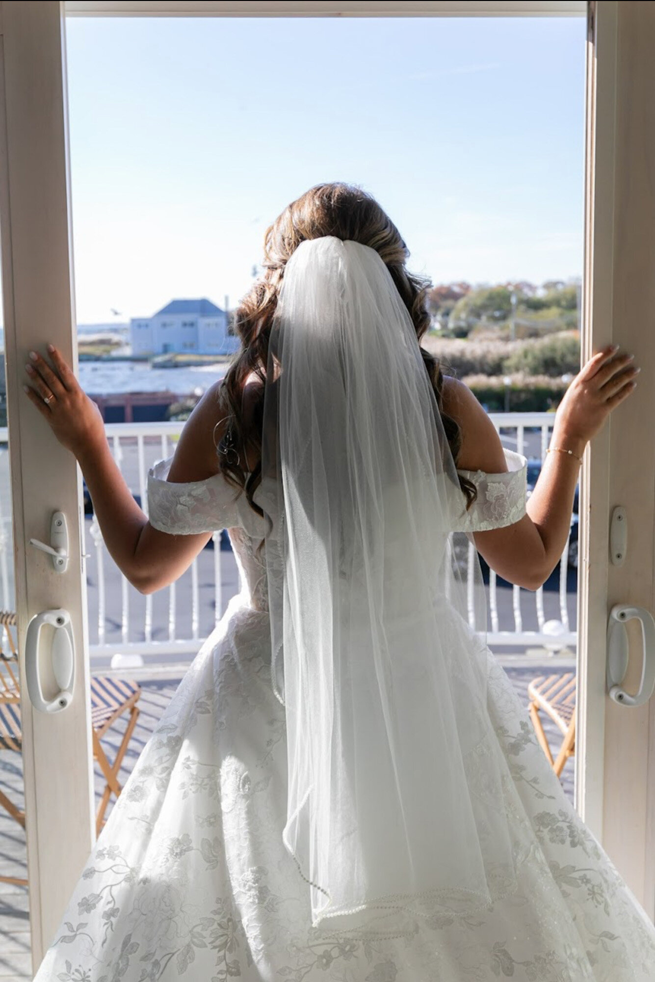 bride in a white off-the-shoulder wedding dress and veil stands with her back to the camera, looking out a set of glass doors onto a sunny outdoor scene with buildings and trees in the distance