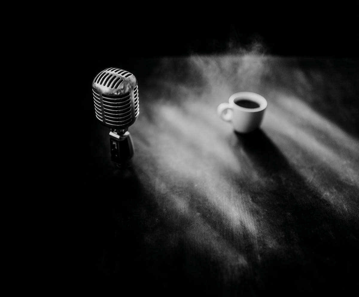 Black-and-white image of a vintage microphone and steaming coffee cup lit by cinematic light beams, symbolizing a poetic and luxurious podcast atmosphere by Jasmina Kimova.