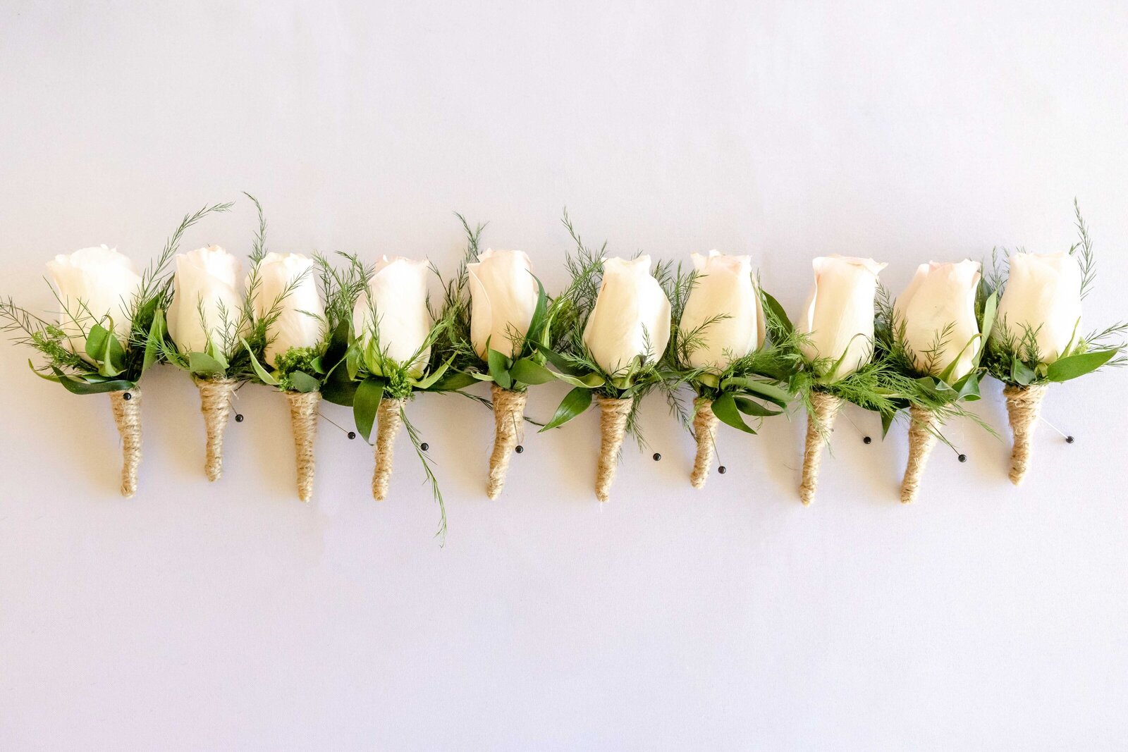 Elegant row of white rose boutonnieres with greenery, prepared for groomsmen, showcasing attention to detail for a Oak Island, NC wedding.