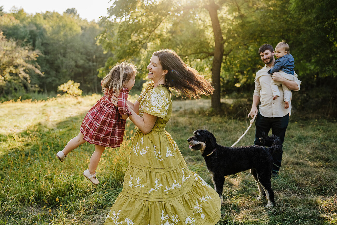 mom twirls daughter around in golden light while dad dog and son watch