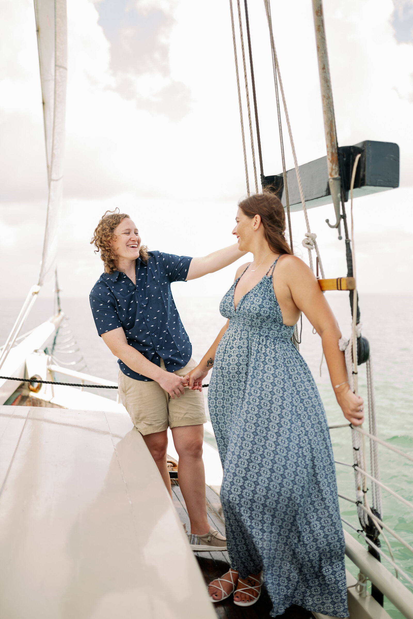 engaged lesbian couple on a sailboat standing together, holding hands, one woman is leaning slightly sideways, both are looking at each other