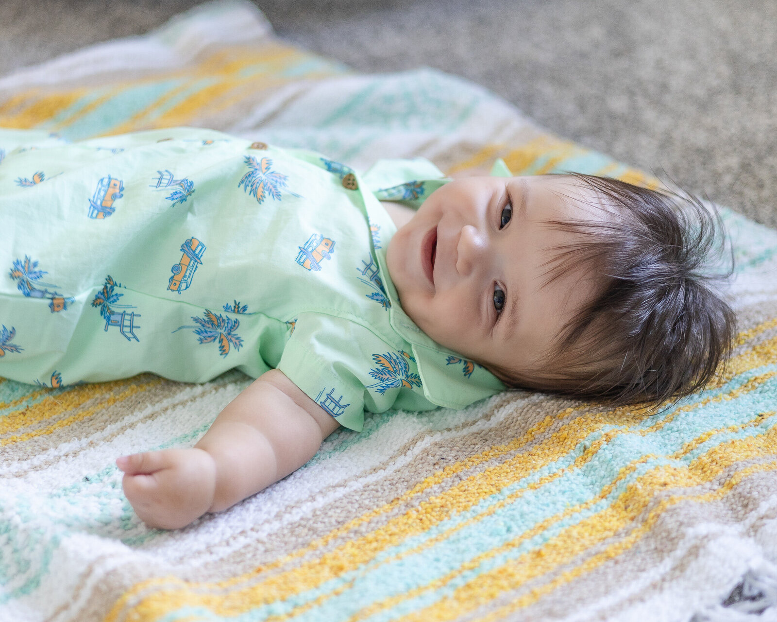 A 6 month old baby with dark brown hair, laying on a blanket, looking at the camera smiling, wearing a light green patterned shirt.