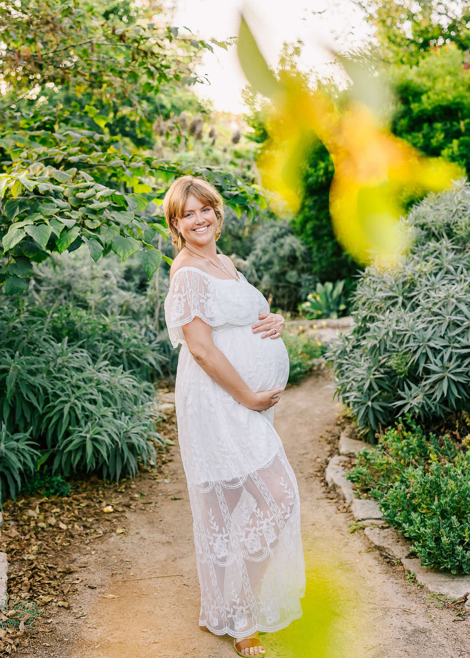 Mama standing smiling holding her baby bump wearing white dress in Huntington Beach, CA by Ashley Nicole.