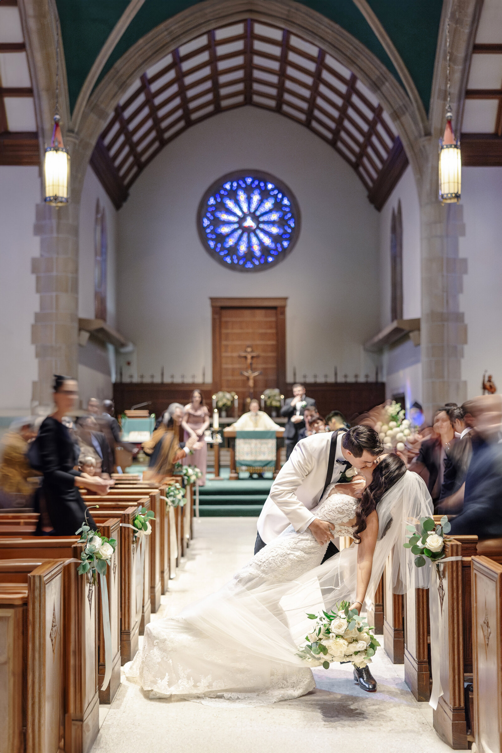 A newlywed couple shares a kiss in the aisle of a church, with the groom dipping the bride. They are surrounded by blurred guests. The church has wooden pews and a large, colorful stained glass window on the wall in the background.