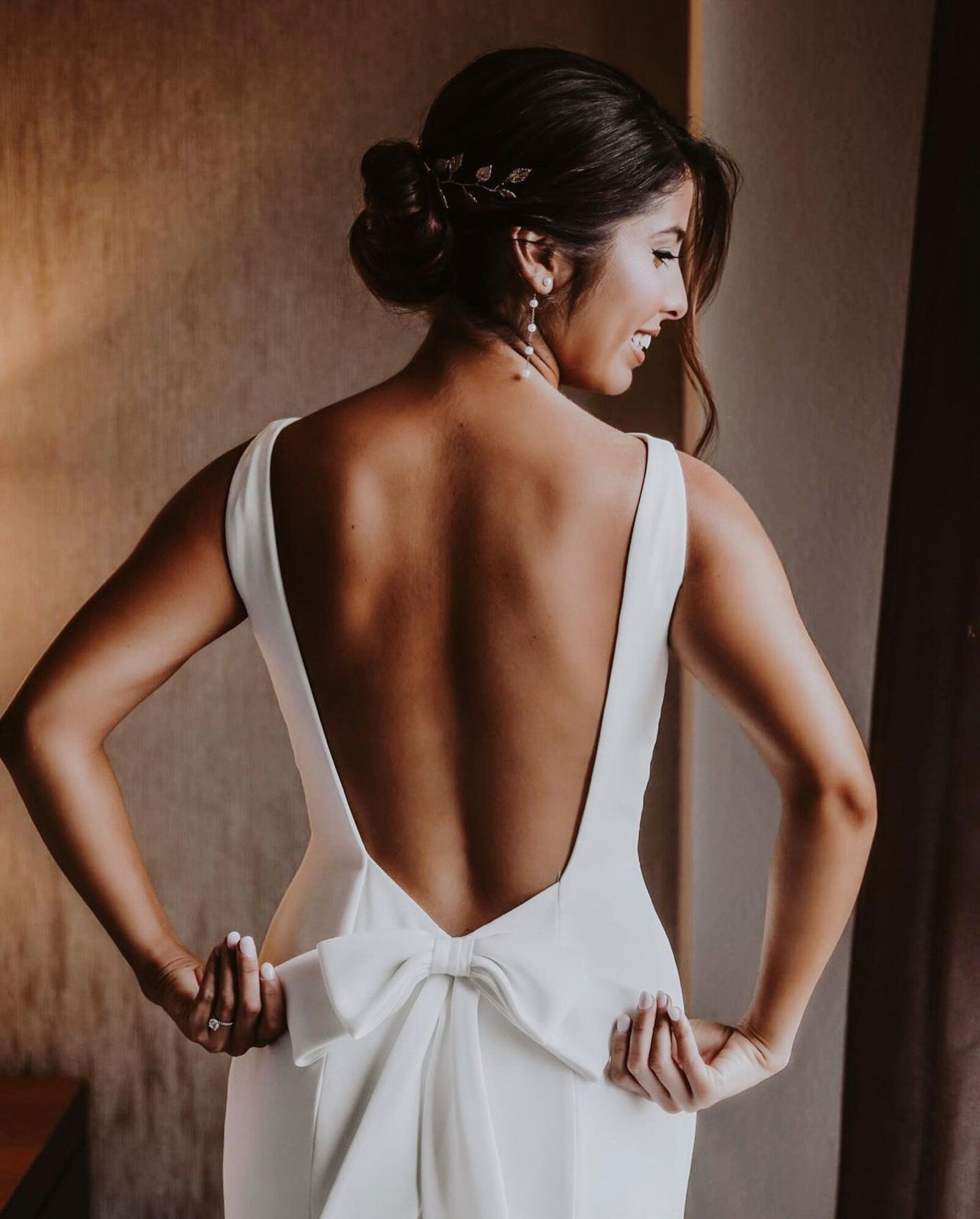 bride standsing with her back to the camera, showcasing an elegant, white backless dress with a large bow at the lower back