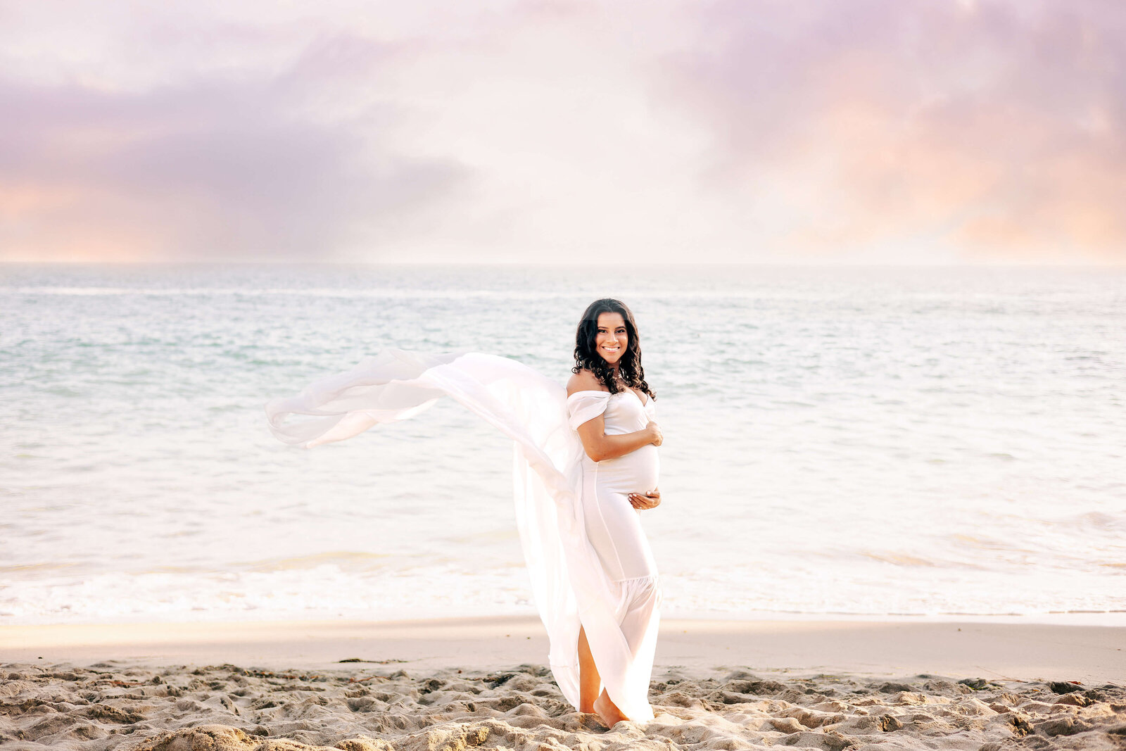 Expectant Mom wearing white dress at beach maternity session with cotton candy skies by Ashley Nicole Photography.