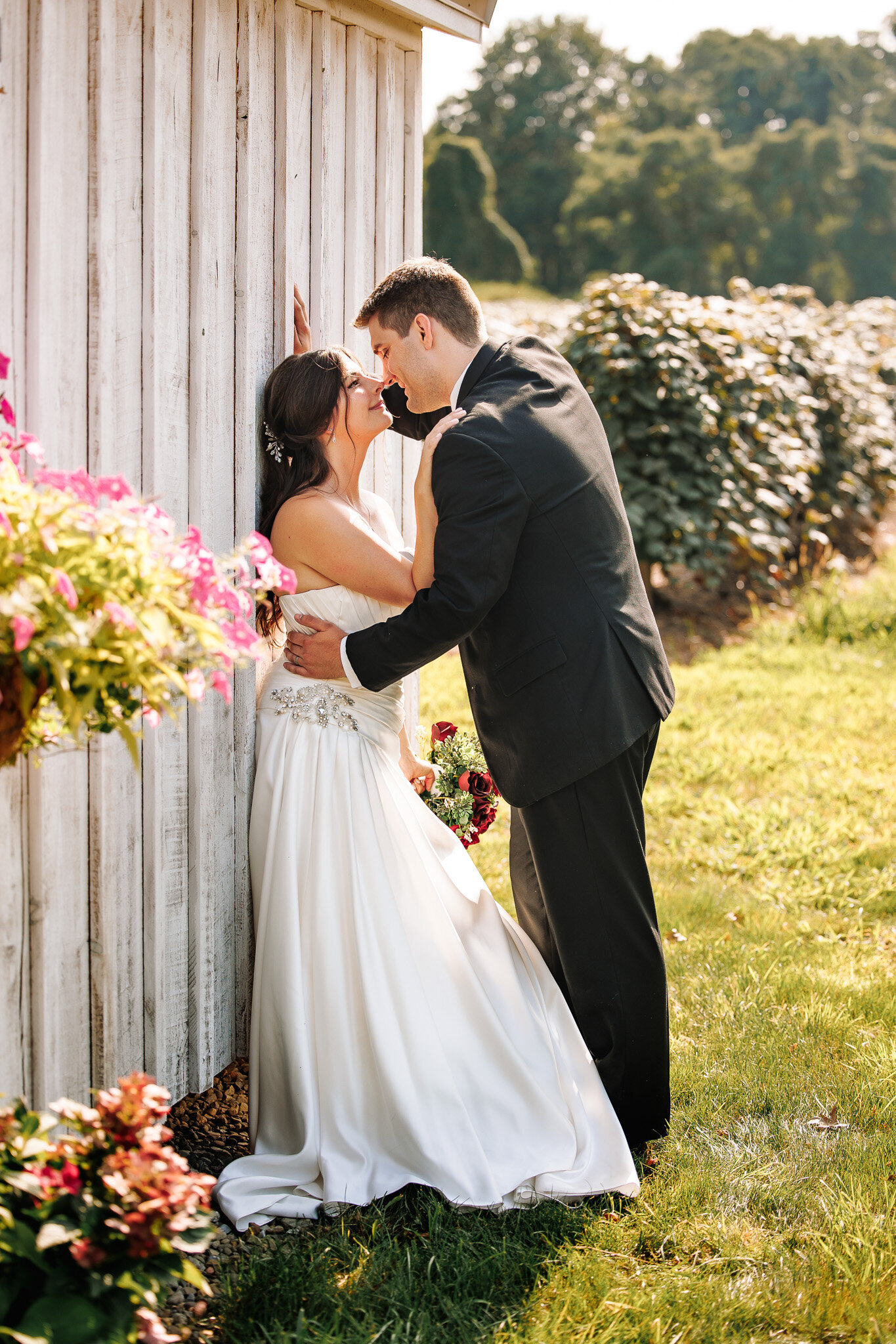 A groom leans his bride up against a white barn going in for a tender kiss. A vineyard in the background.
