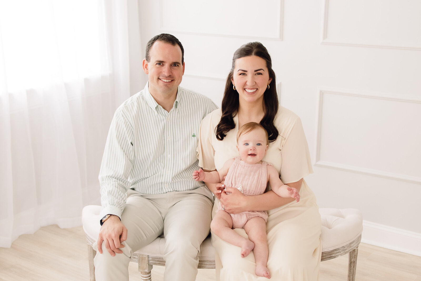 Sweet family wearing light, neutral clothing and all smiling at the camera in a bright studio in Louisville