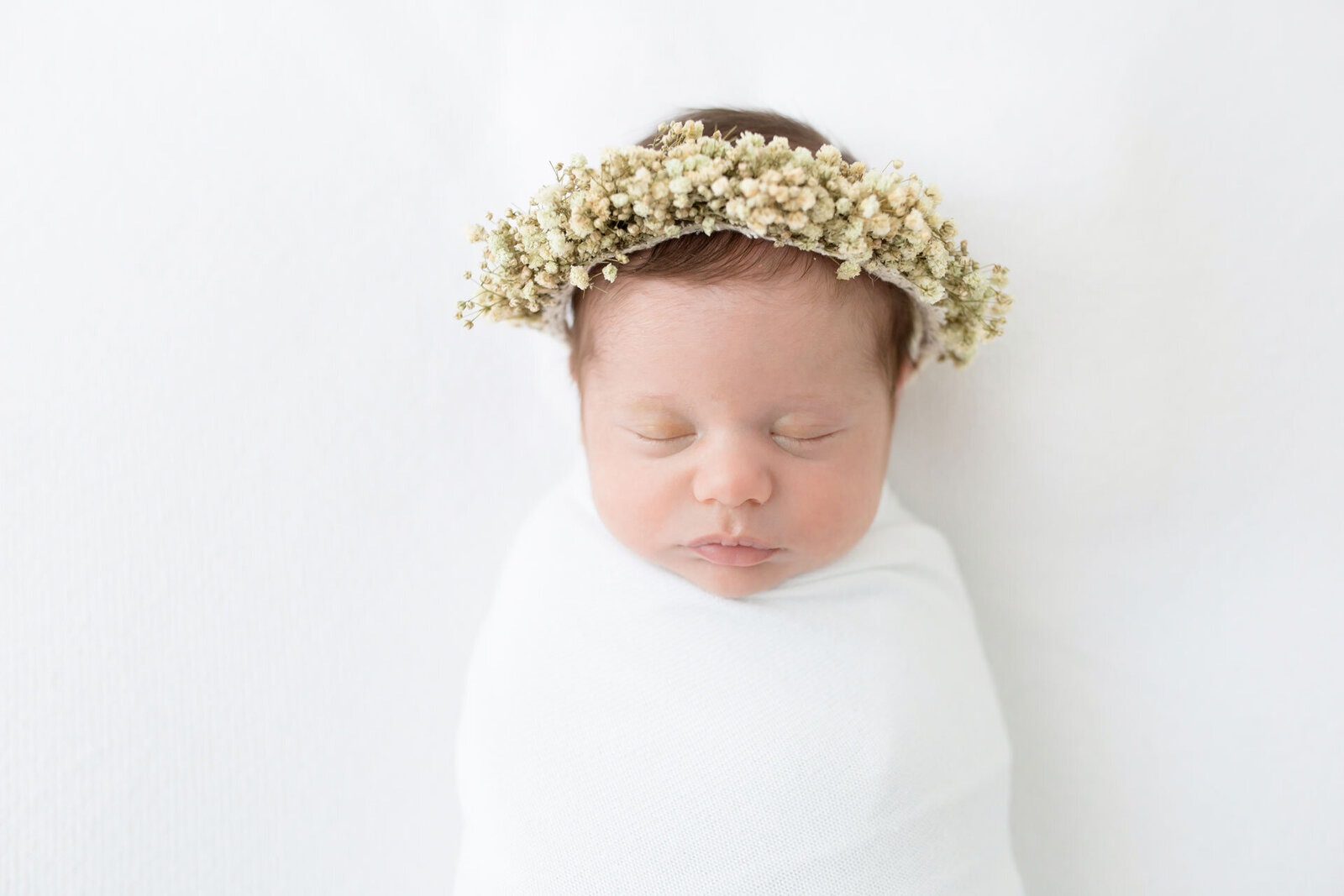 Neugeborenenshooting Bielefeld: Schlafendes Baby in weißer Decke mit üppigem Blumenkranz auf dem Kopf