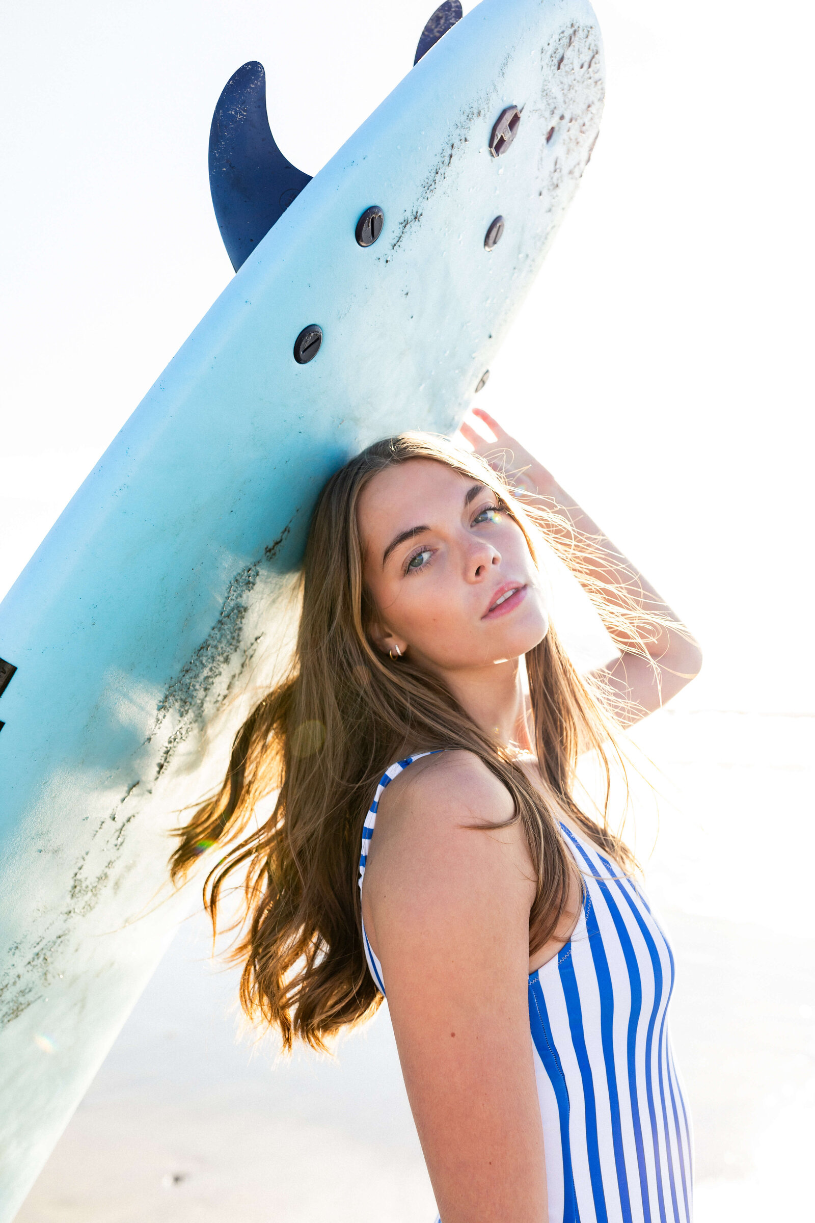 brunette girl holding a surf board wearing a striped one piece swimsuit