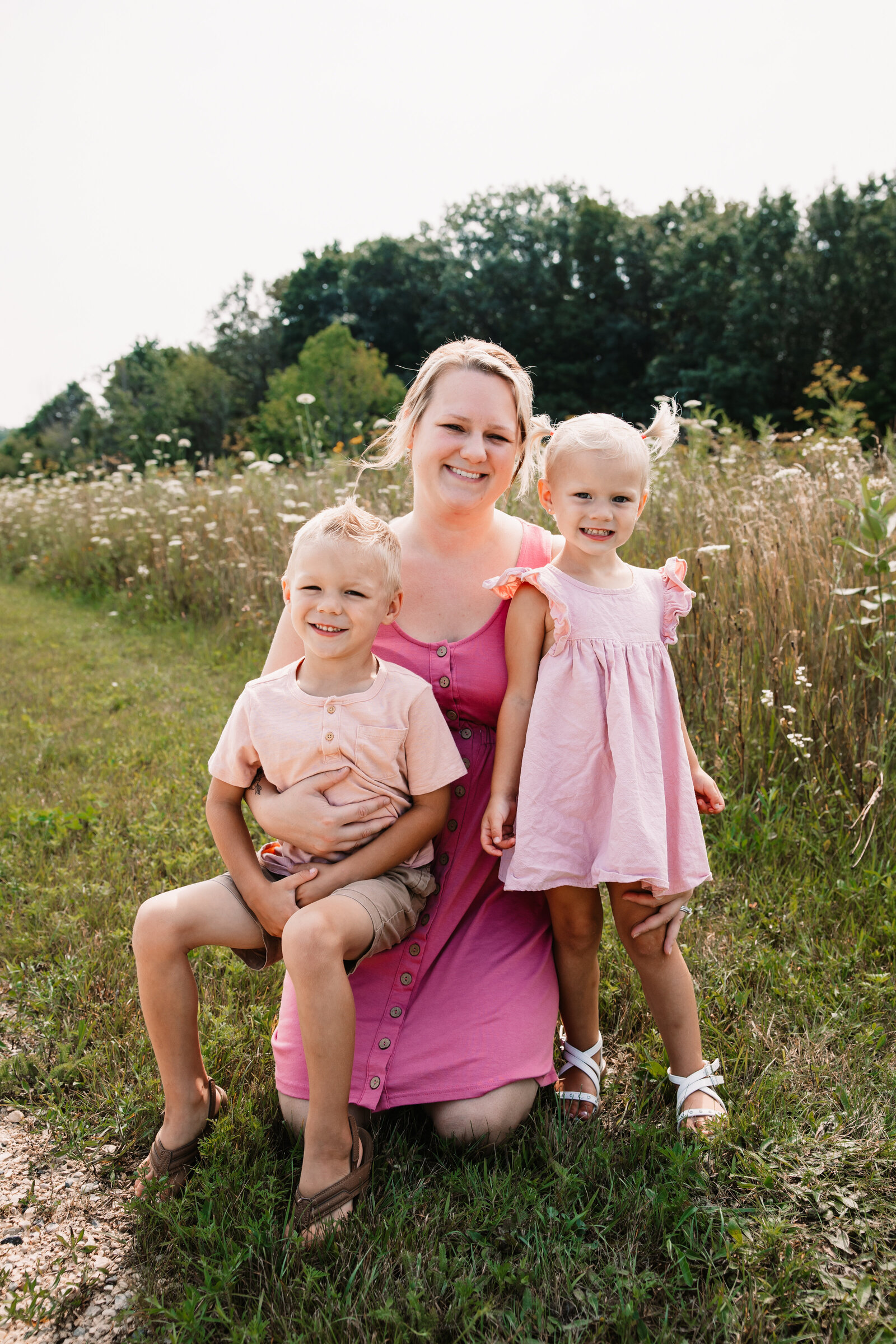 A smiling mom in a pink dress kneels in a park trail with her toddler son and daughter both in pink