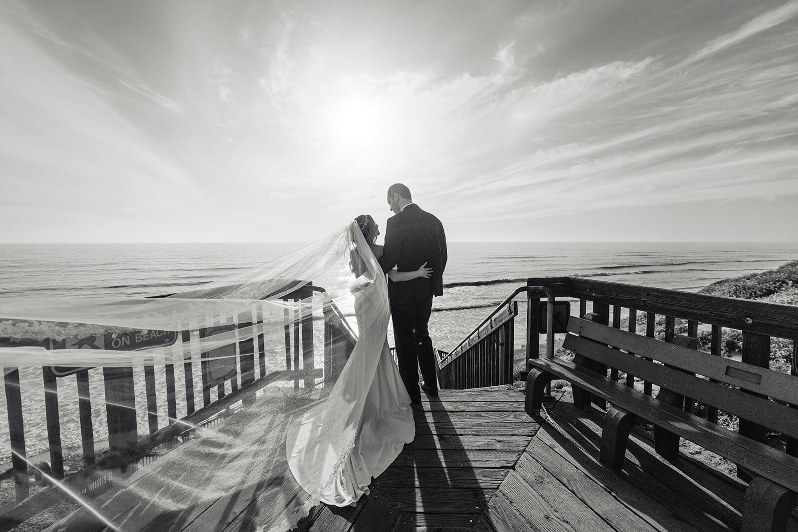 black and white photo of bride and groom on a deck facing the ocean with the bride's veil blowing in the wind