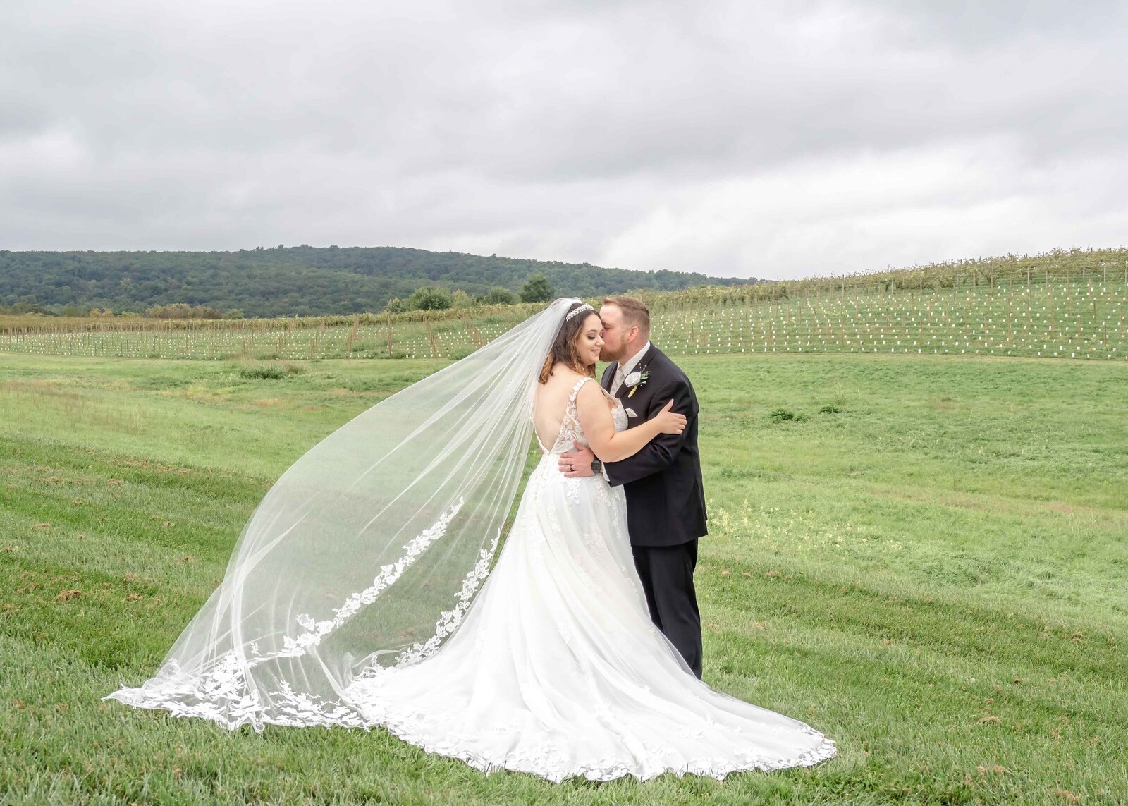 The bride and groom share a romantic kiss in a lush green meadow with rolling hills and mountains in the background, captured during a Boone, NC wedding, blending rustic elegance with natural beauty.